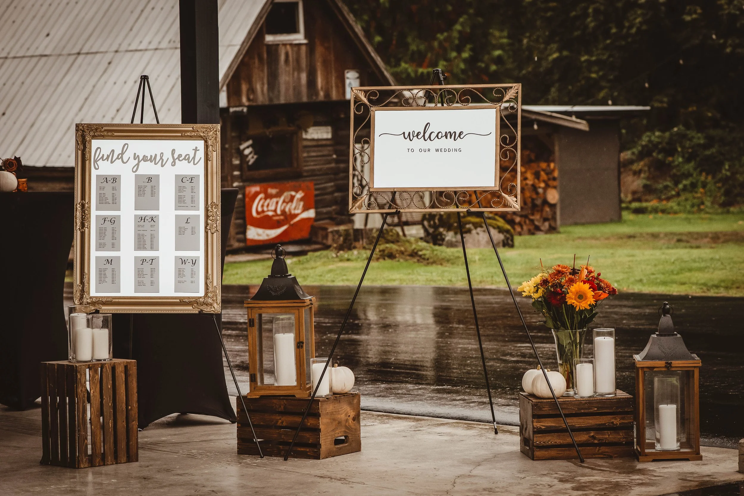 Wedding welcome sign, seating chart, candles, flowers, and lanterns outdoors on a rainy day.