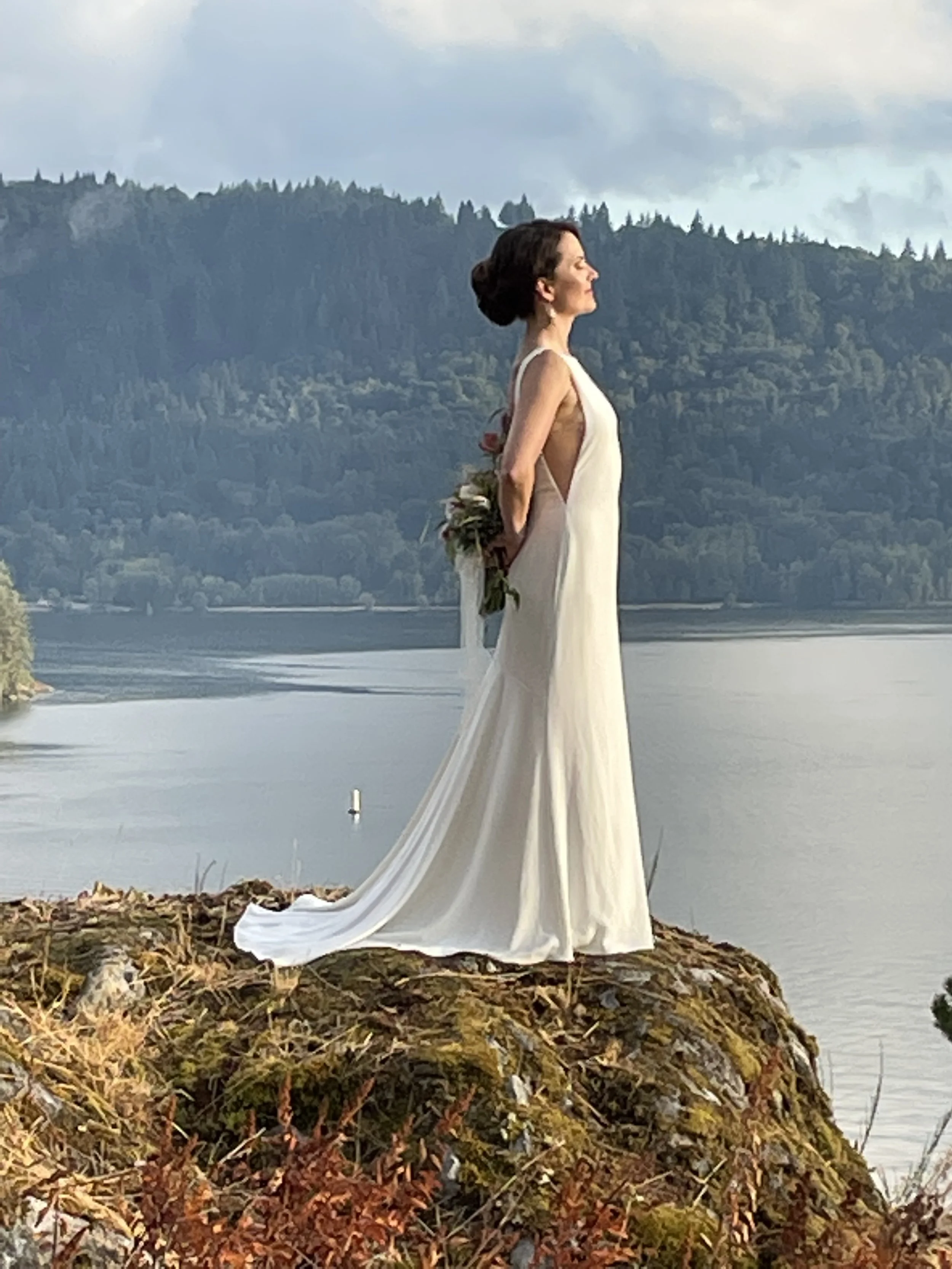 A woman in a white dress standing on a rocky outcrop beside a body of water, holding a bouquet behind her back, with forested hills in the background.