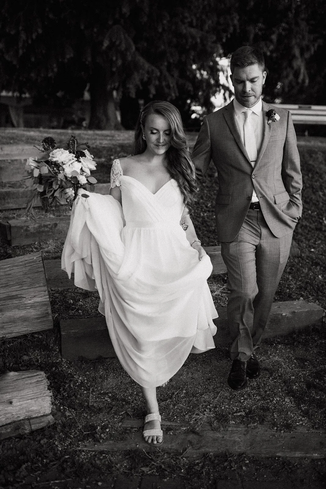 Black and white photo of a bride and groom walking outdoors. The bride is in a white wedding dress, holding a bouquet of flowers, and lifting her dress slightly. The groom wears a suit with a tie, walking with hands in pockets. They are on a dirt path with trees and steps in the background.