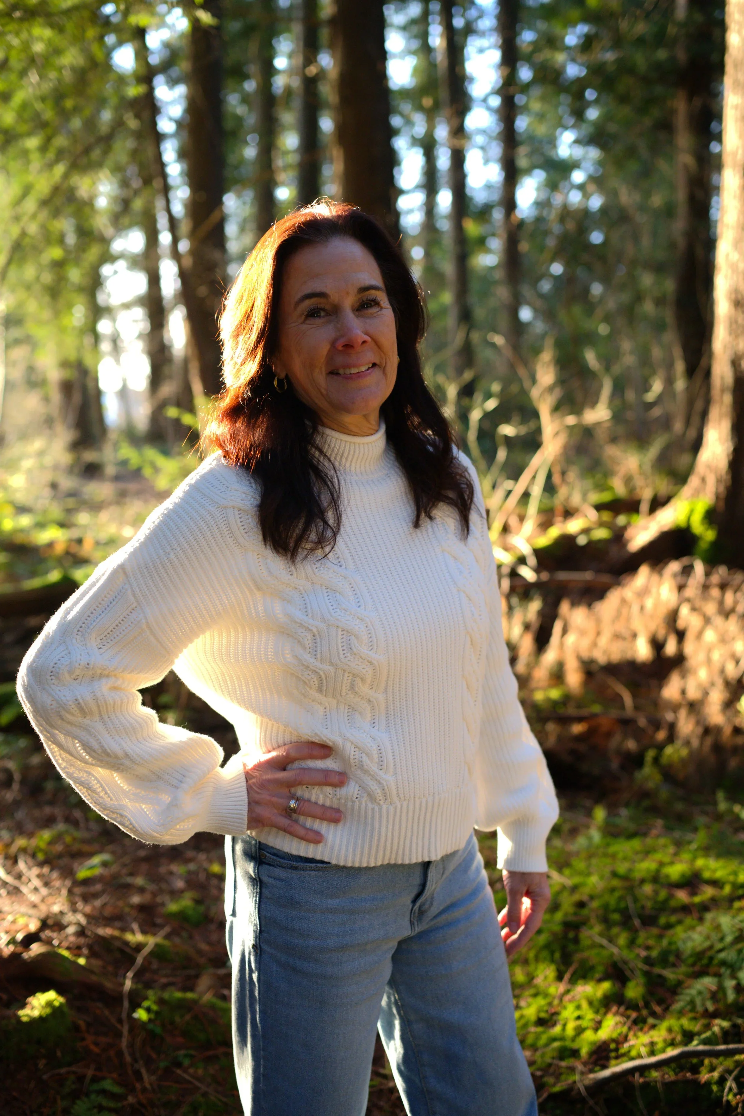 Woman with dark hair in a white sweater and blue jeans standing in a forest with sunlight filtering through trees.