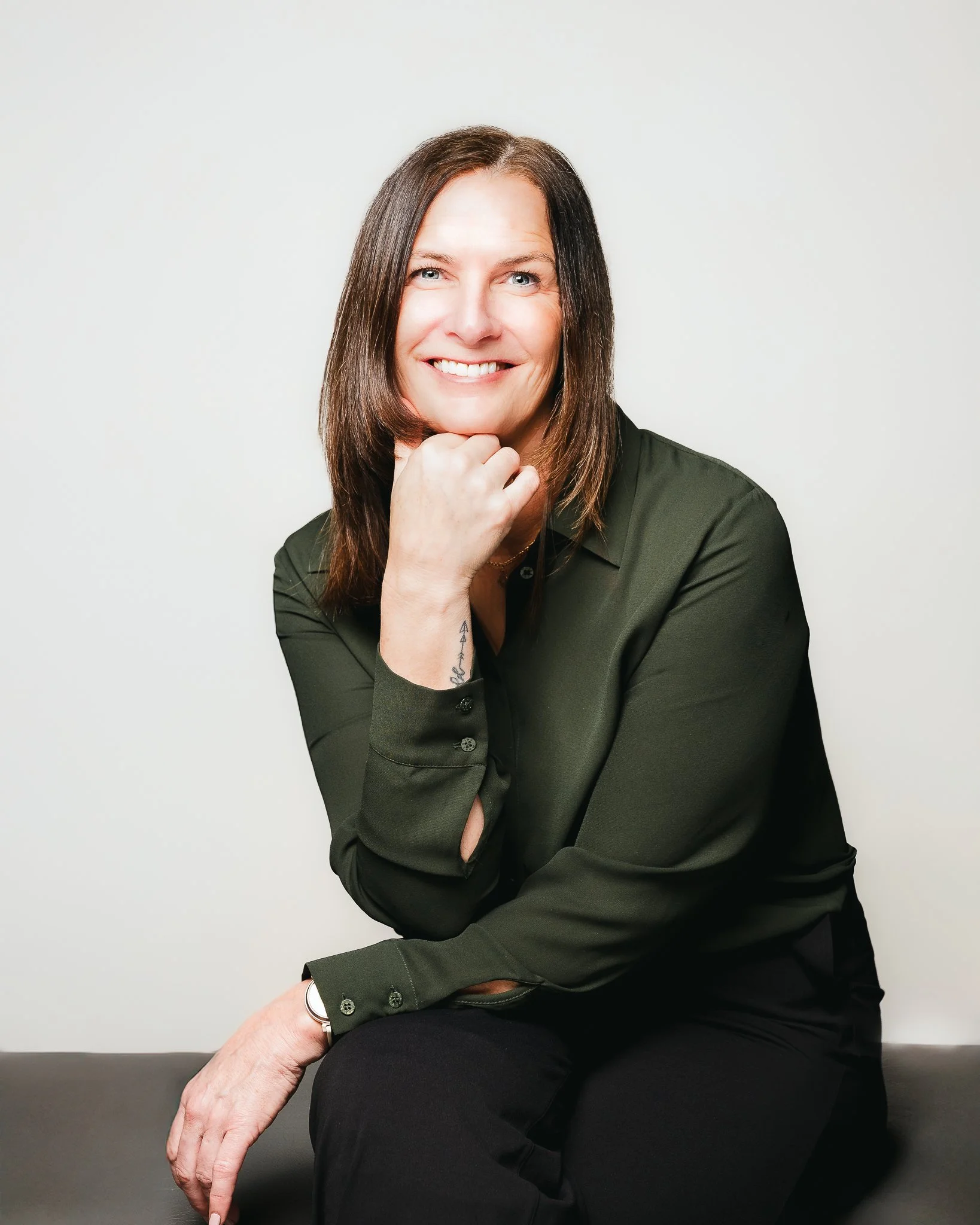 A smiling woman with brown hair, wearing a dark green blouse, sitting with her chin resting on her hand, against a plain light-colored background.