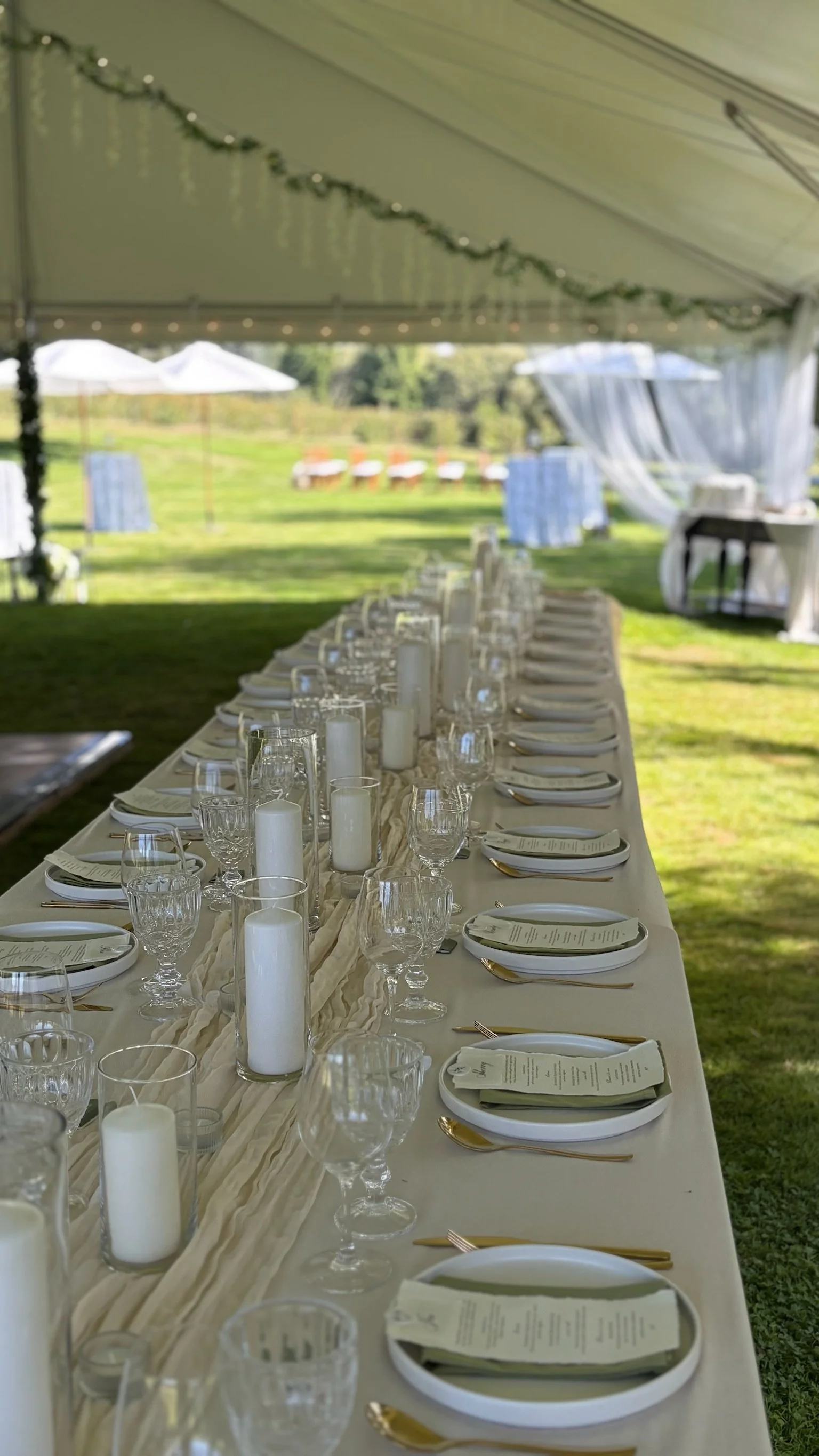 Long dining table set for an outdoor event under a tent, with white plates, gold utensils, clear glasses, and white candles, in a grassy yard with umbrellas and trees in the background.