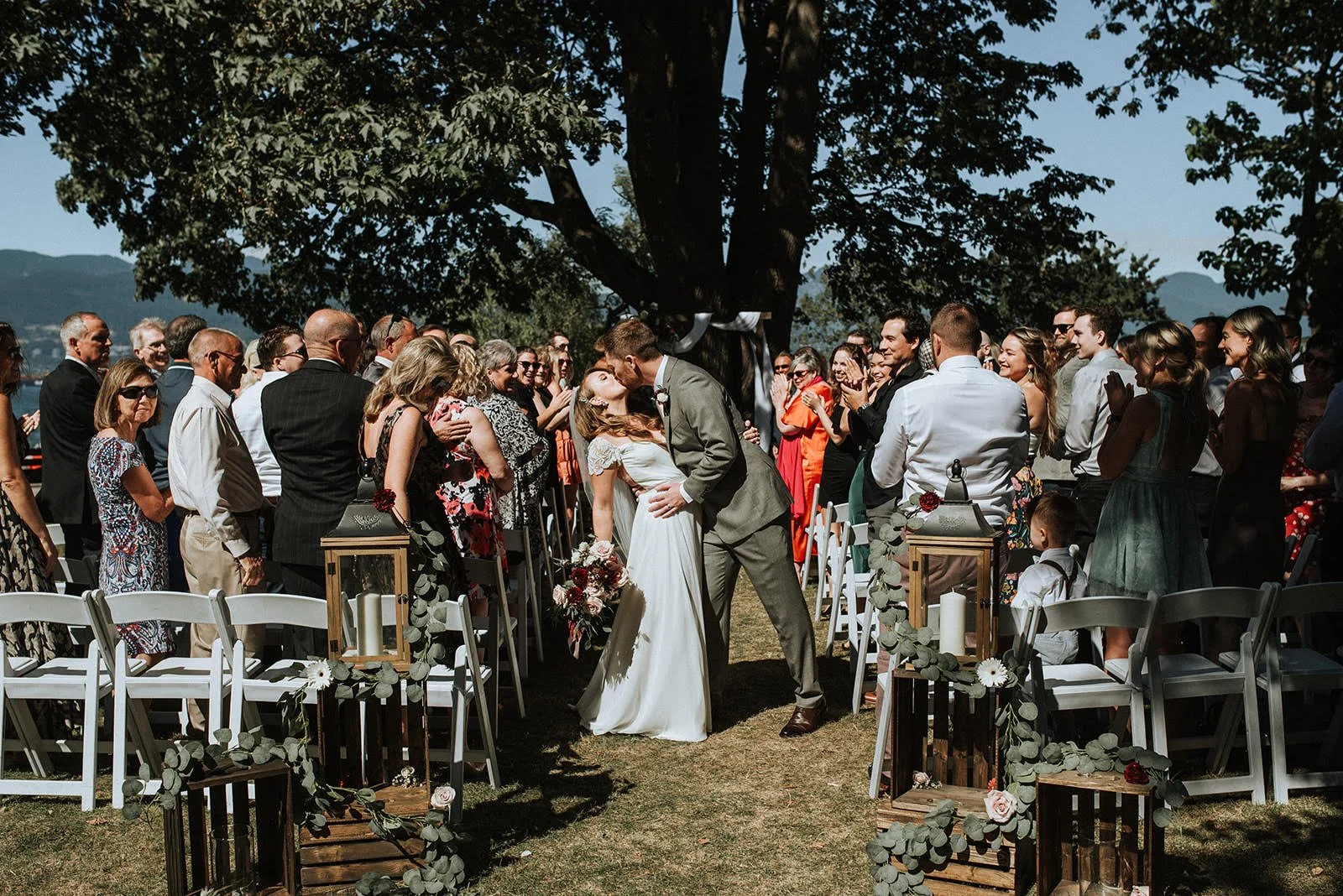 A bride and groom kiss at their outdoor wedding ceremony surrounded by guests.