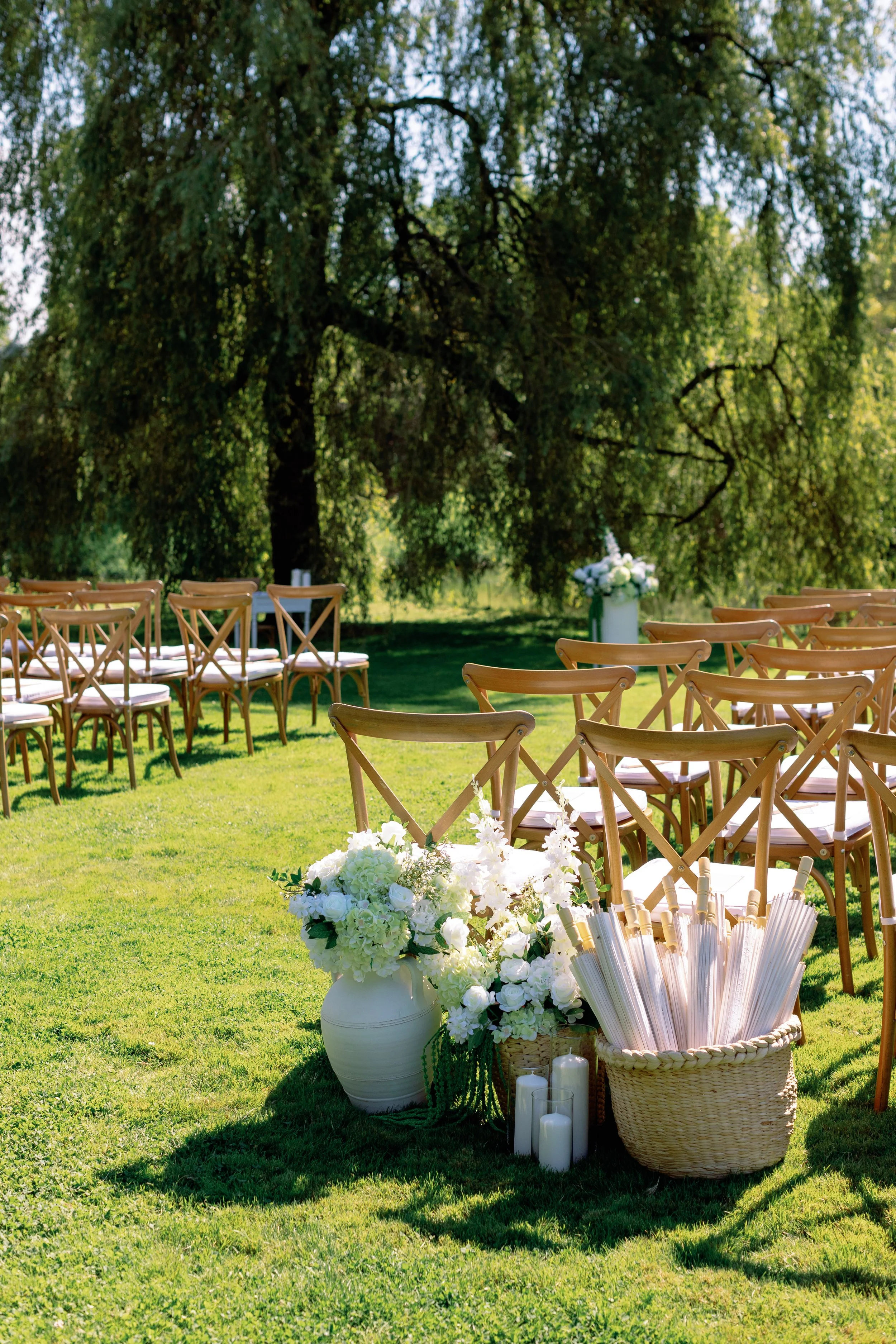 Outdoor wedding ceremony setup with rows of wooden chairs, floral arrangements, candles, and a large tree in the background.