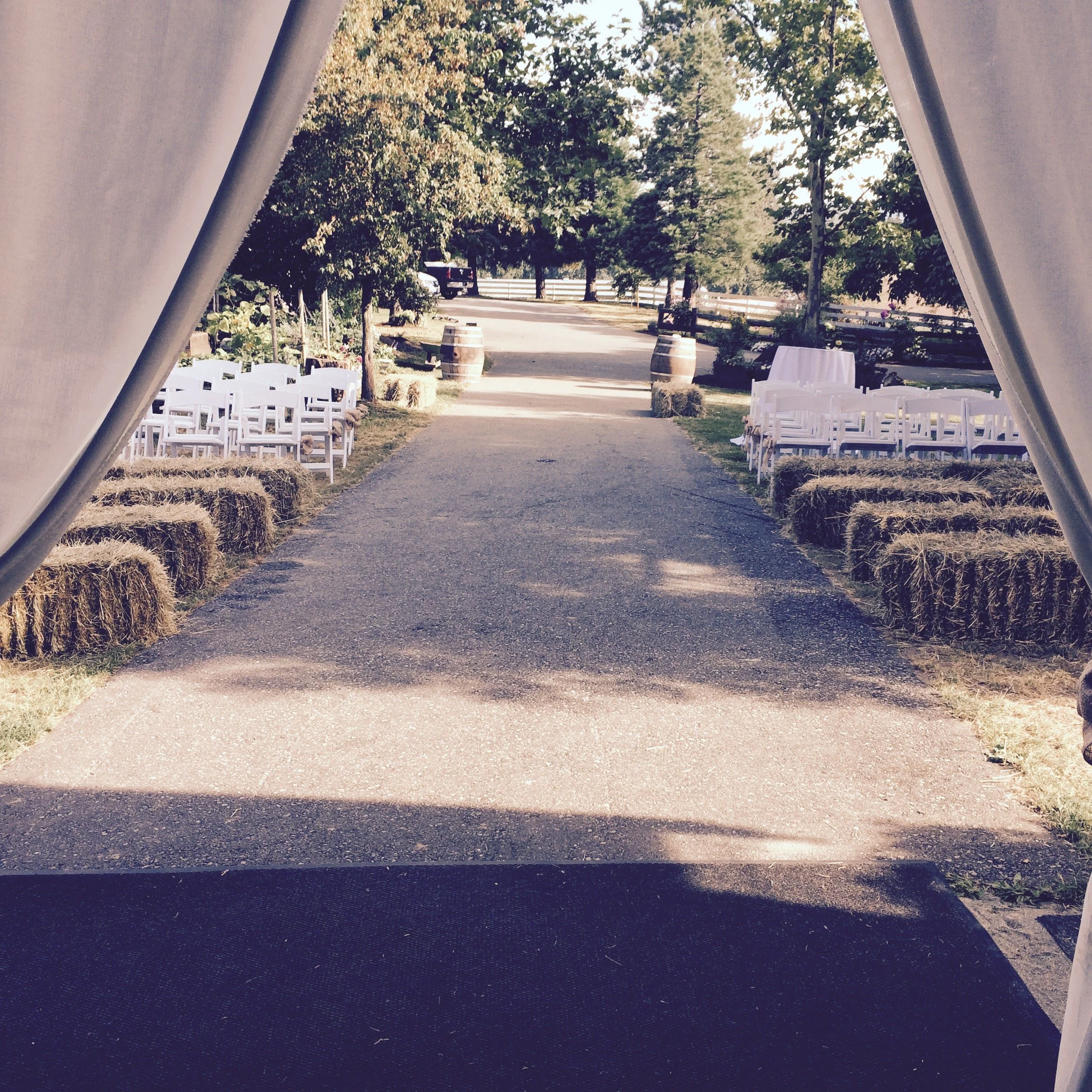 Outdoor wedding ceremony setup with hay bales and white chairs on either side of a central aisle, viewed from inside a tent.