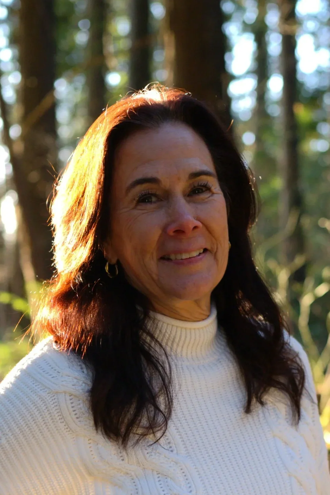 A woman with long dark hair stands outdoors in a forest, smiling with the sunlight illuminating her hair and face.