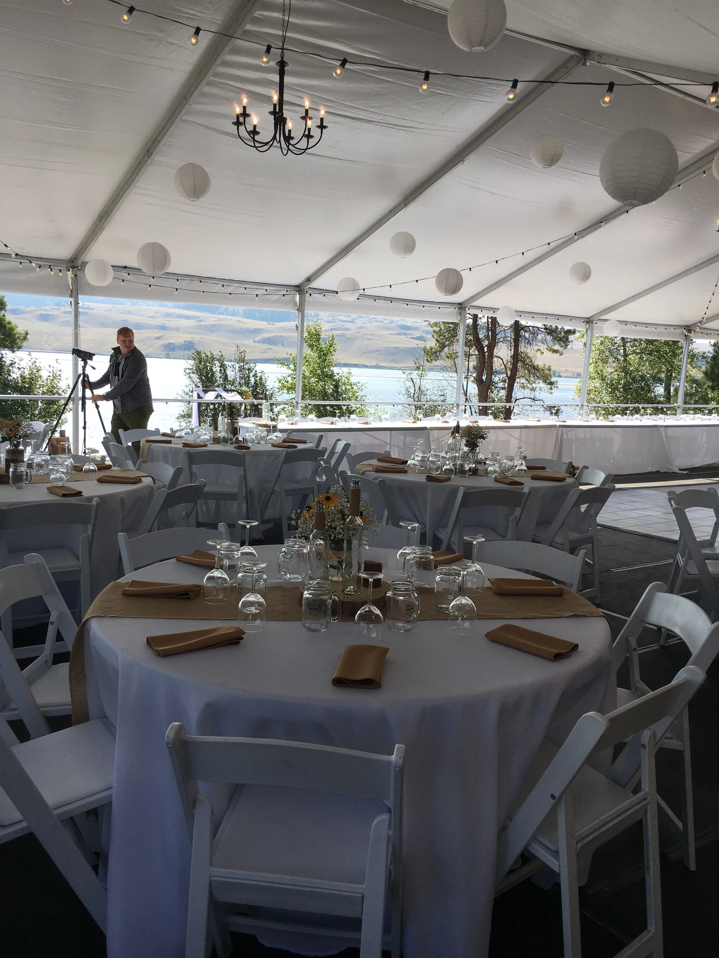 A decorated outdoor event space with tables set for a gathering, featuring white tablecloths, brown napkins, glassware, and centerpieces with flowers. Hanging paper lanterns and string lights decorate the tent. A person is adjusting a camera on a tri