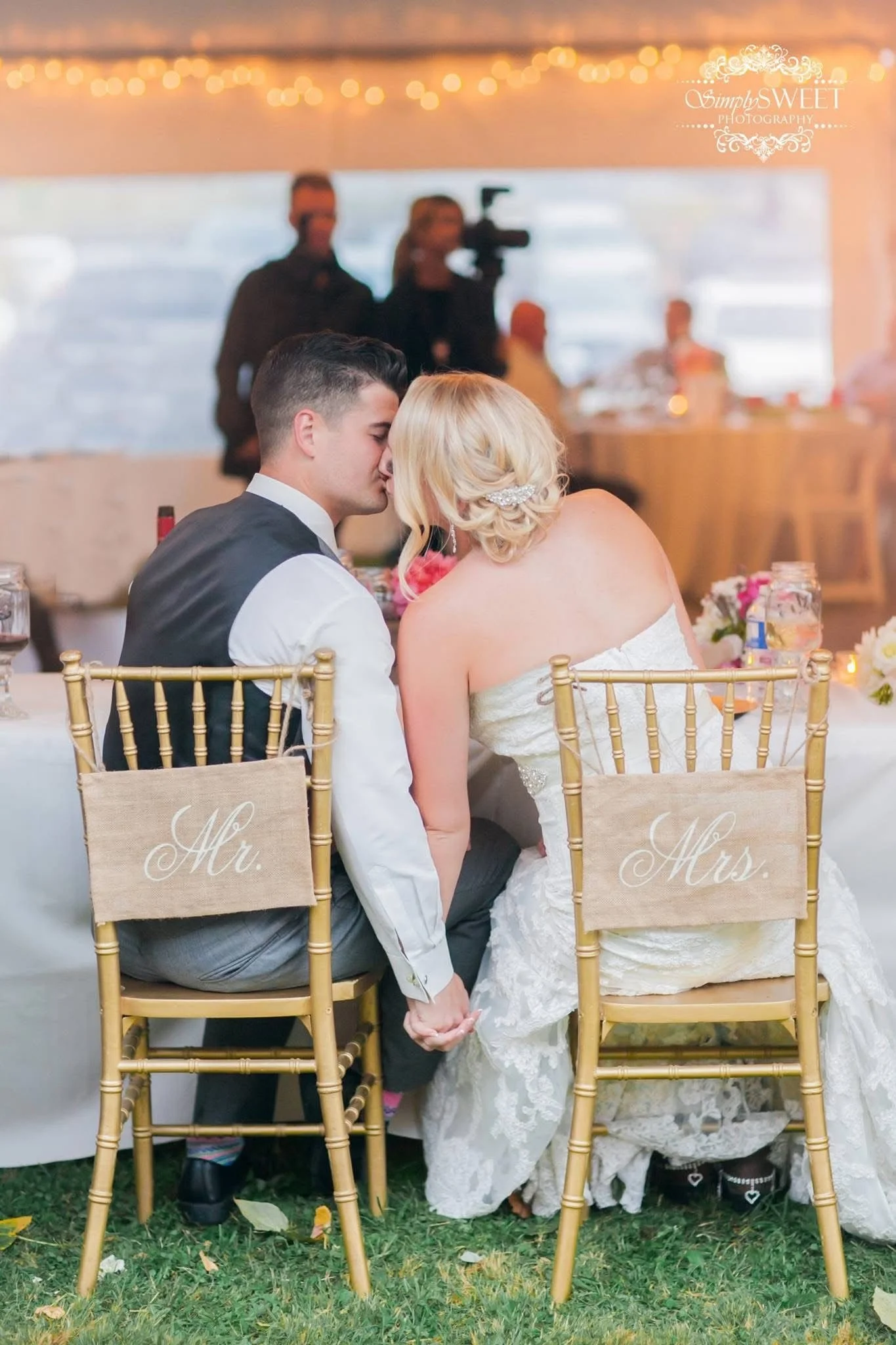 A bride and groom sitting close together, leaning in for a kiss during their wedding reception. They are holding hands under the table, with signs on their chairs reading 'Mrs.' and 'Mr.'.
