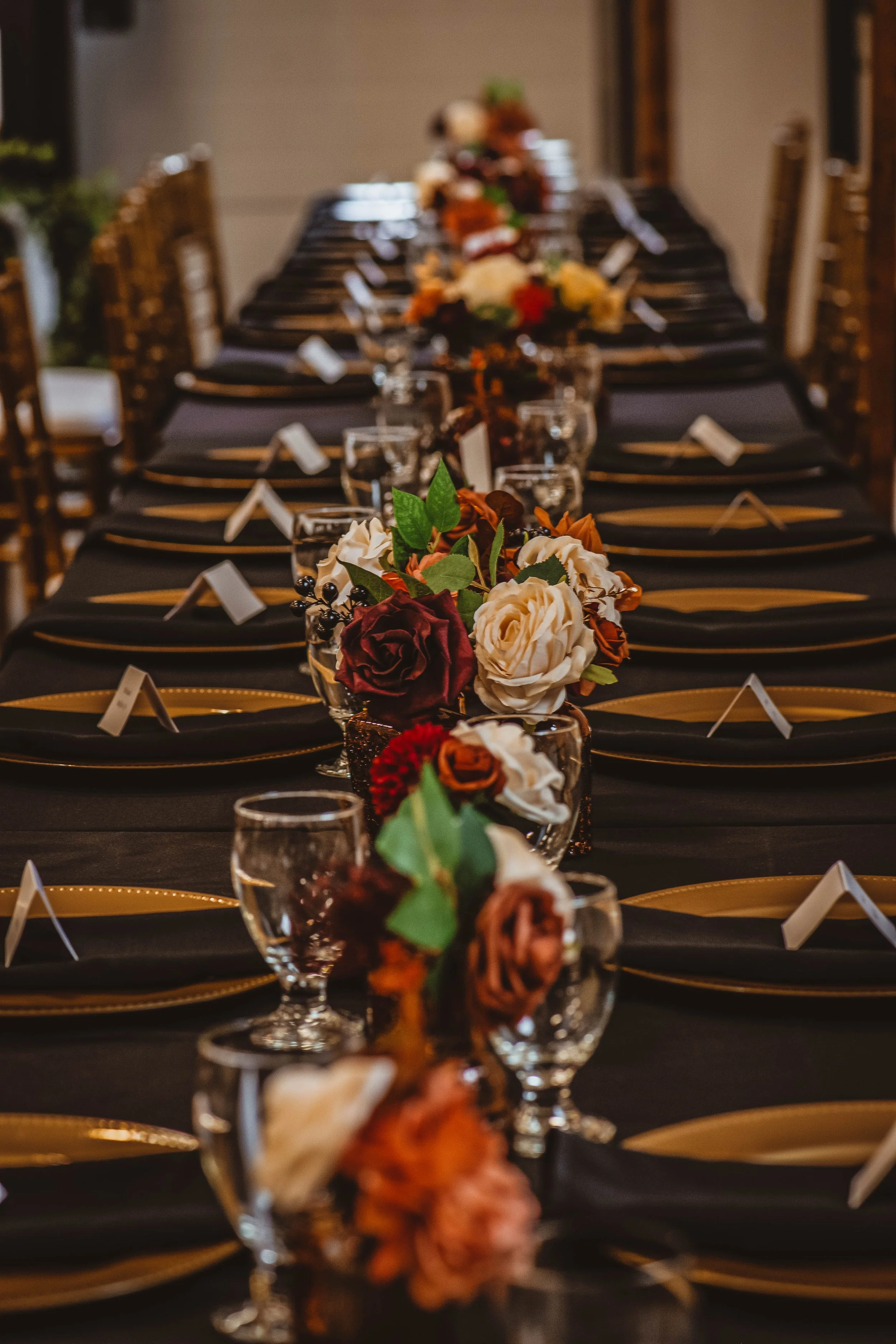 A long banquet table set for a formal event, decorated with black tablecloths, gold-accented black plates, clear wine glasses, and floral centerpieces featuring white, red, and blush roses, along with greenery.