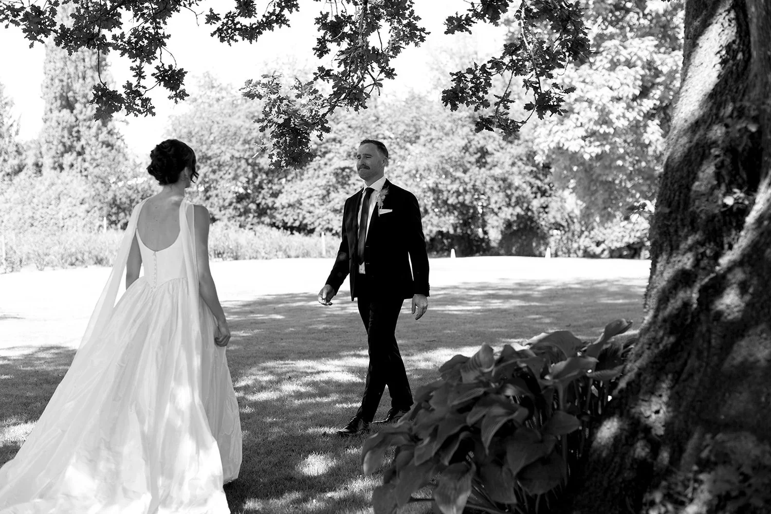 A black-and-white photo of a bride in a wedding dress and a groom in a suit standing outdoors under a tree, with a grassy area and trees in the background.