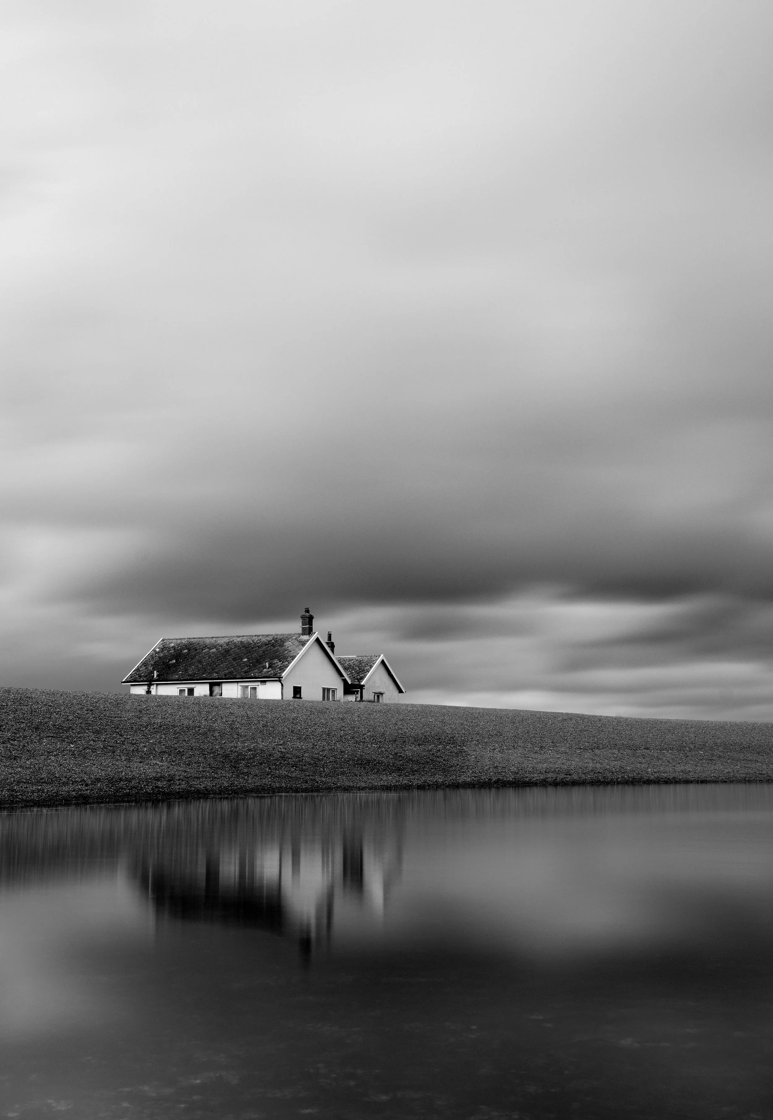 A black-and-white photograph of a house with a chimney on a grassy hillside near a calm body of water, with dark, cloudy skies overhead.