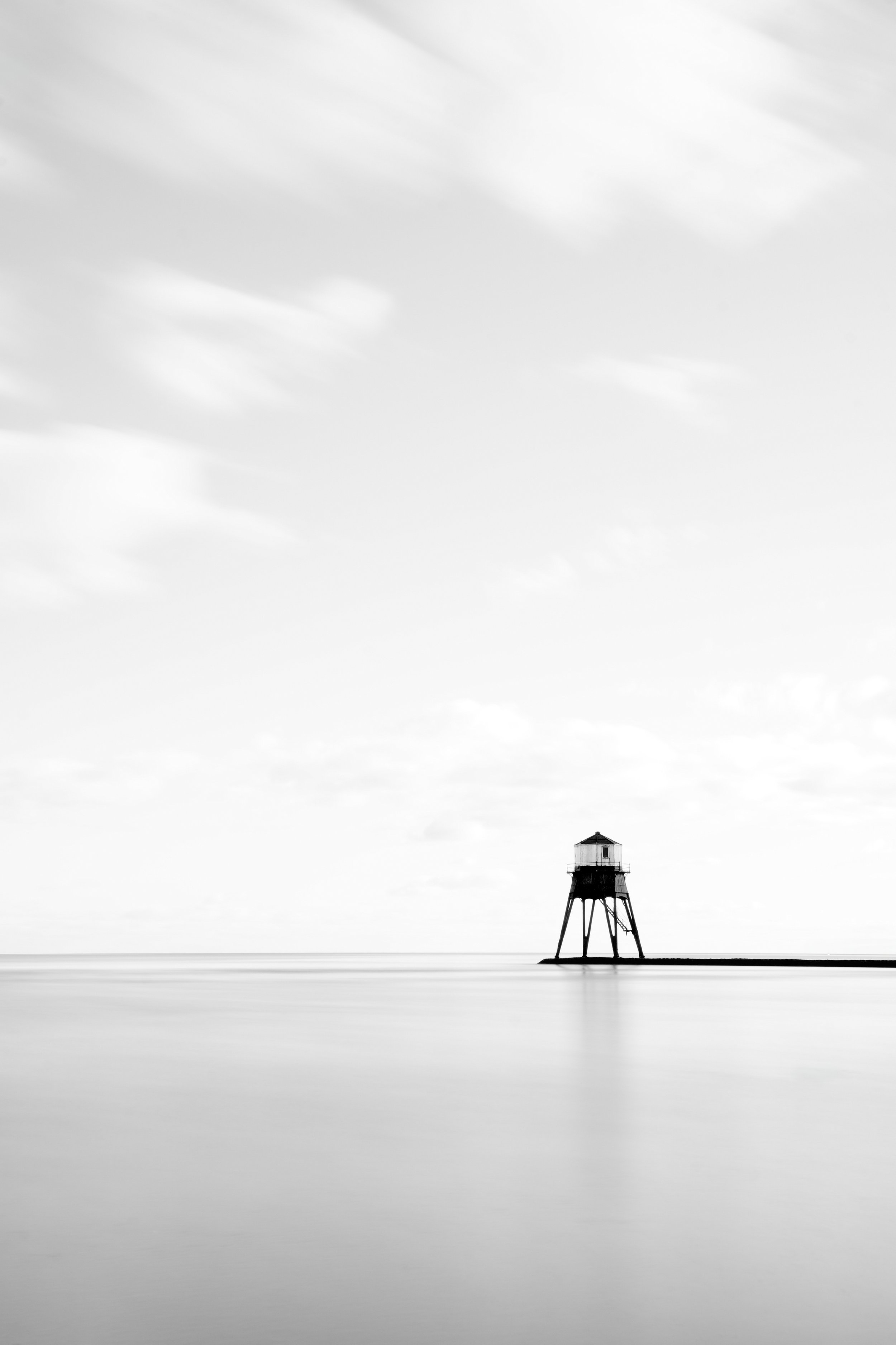 A black-and-white photo of a lone lighthouse on stilts over calm water with a cloudy sky in the background.