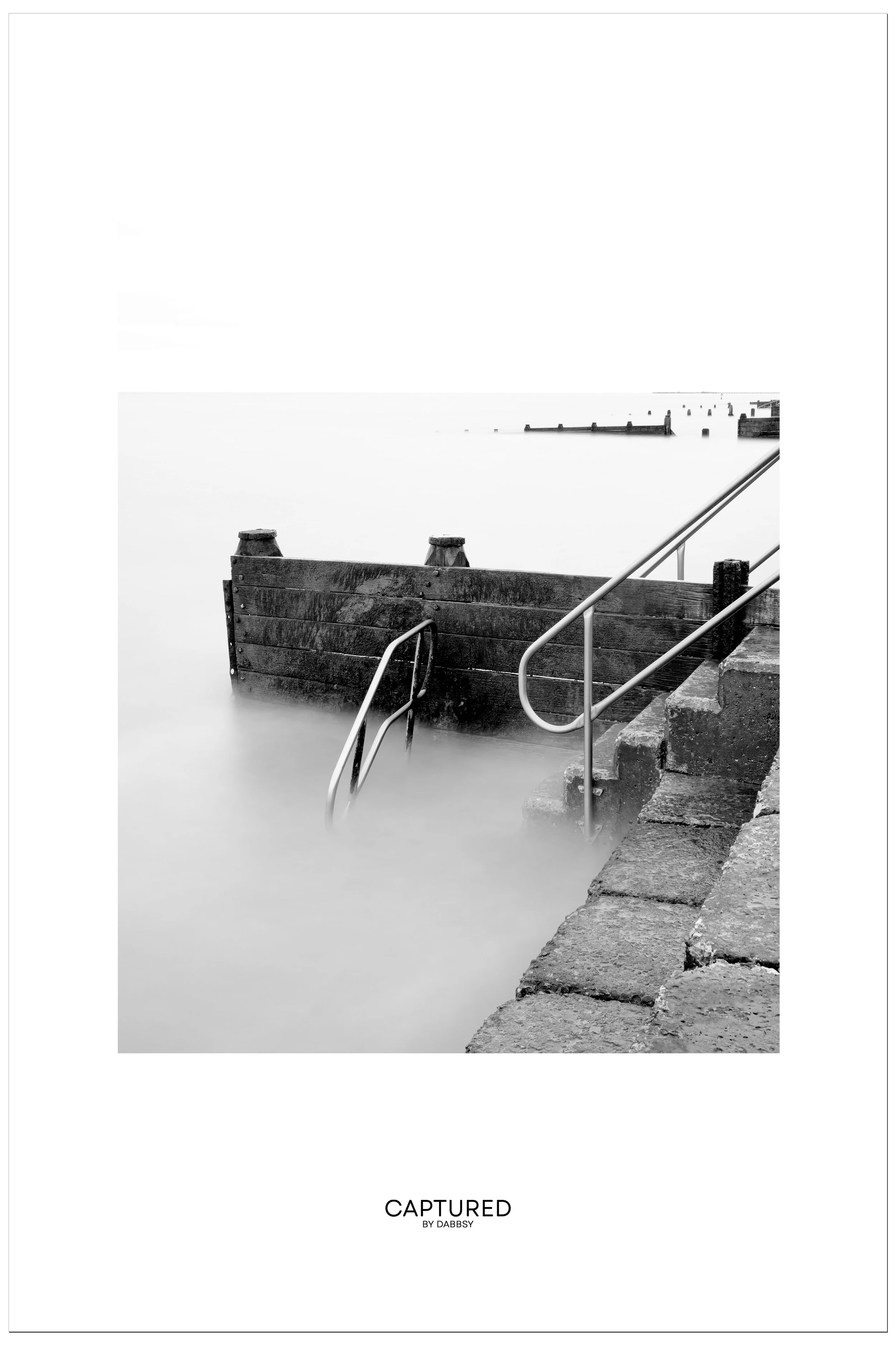 Black and white photo of a concrete and wood pier with metal railing leading into calm water, with distant breakwaters and sky in background, located at Walton on the Naze