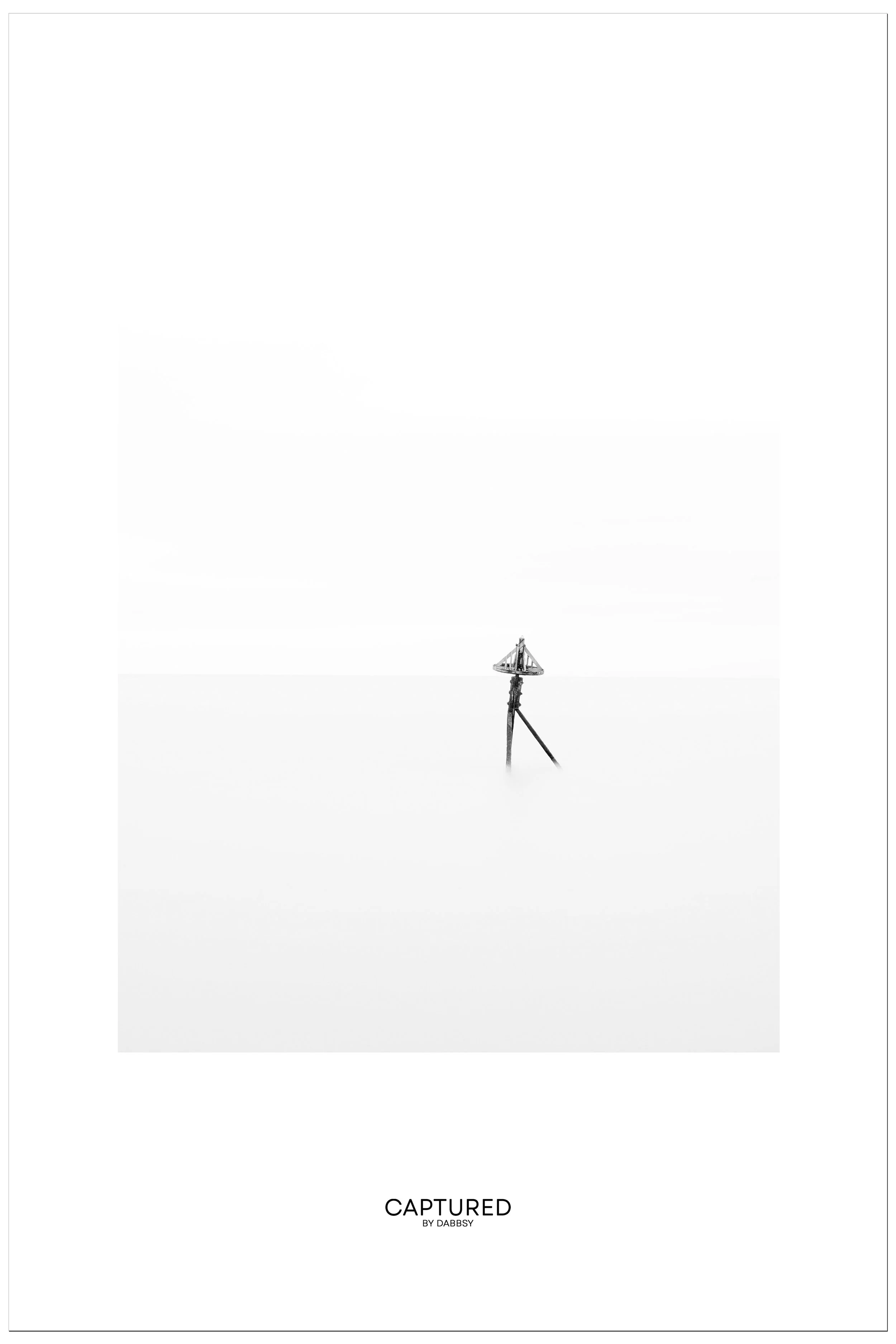 A minimalist black-and-white photograph featuring a compass-like navigation marker on a pole emerging from a calm, foggy sea at Walton on the Naze. The image has a vast, empty sky with no clouds or other objects visible. 