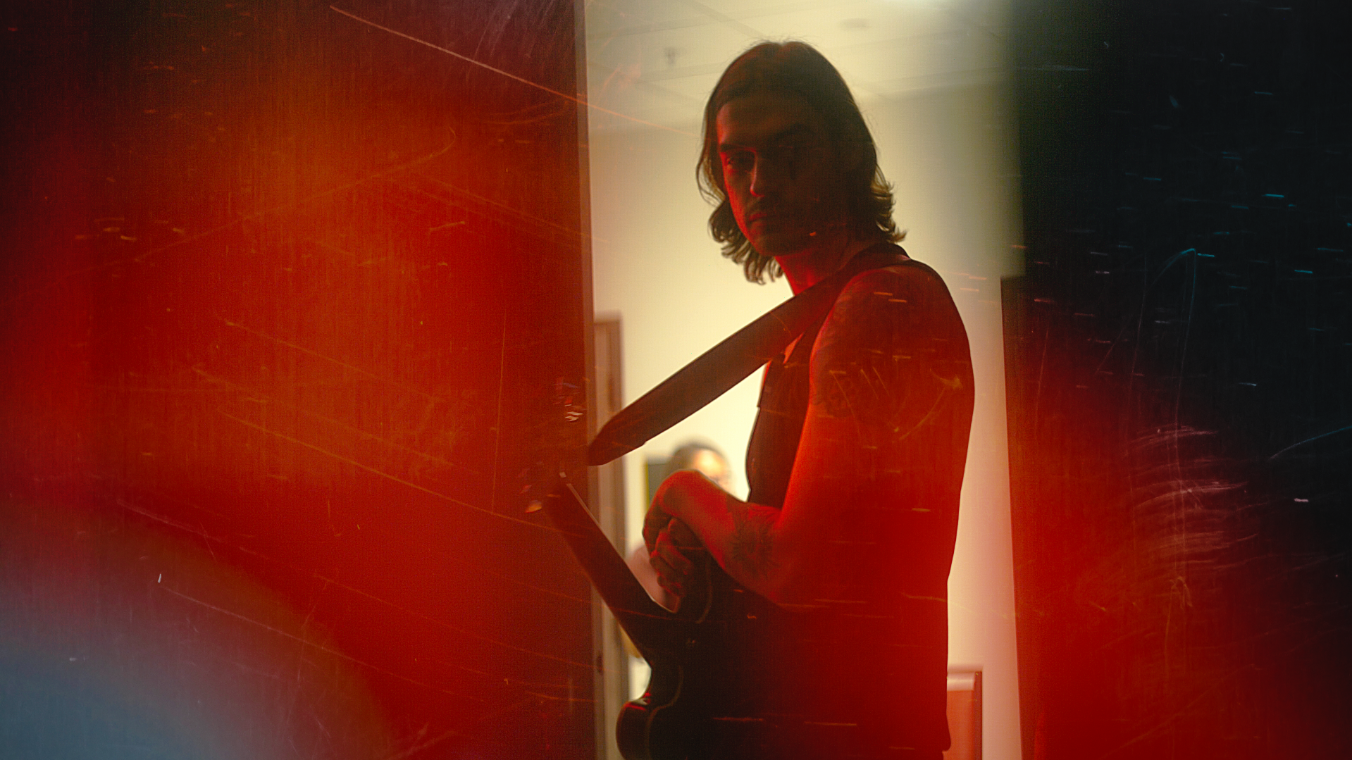 A person with long hair holding a guitar, standing in a dimly lit room with red and dark lighting, facing the camera.