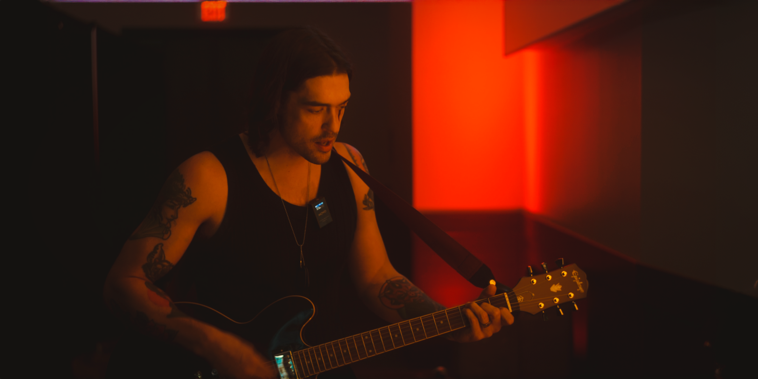 A man with long hair and tattoos on his arms is playing an electric guitar in a dimly lit room with red lighting.
