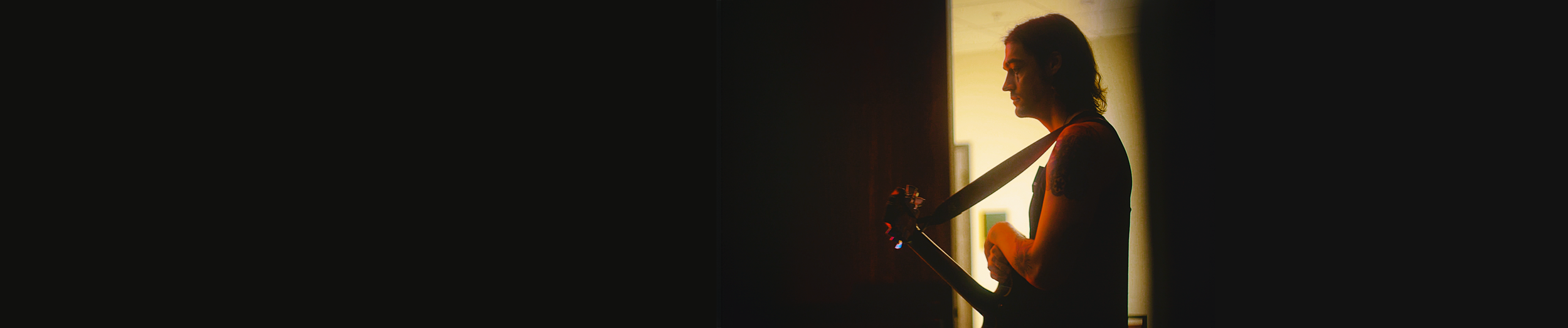 Person with long hair in silhouette, holding an electric guitar in front of a lit doorway, backlit by warm yellow light.