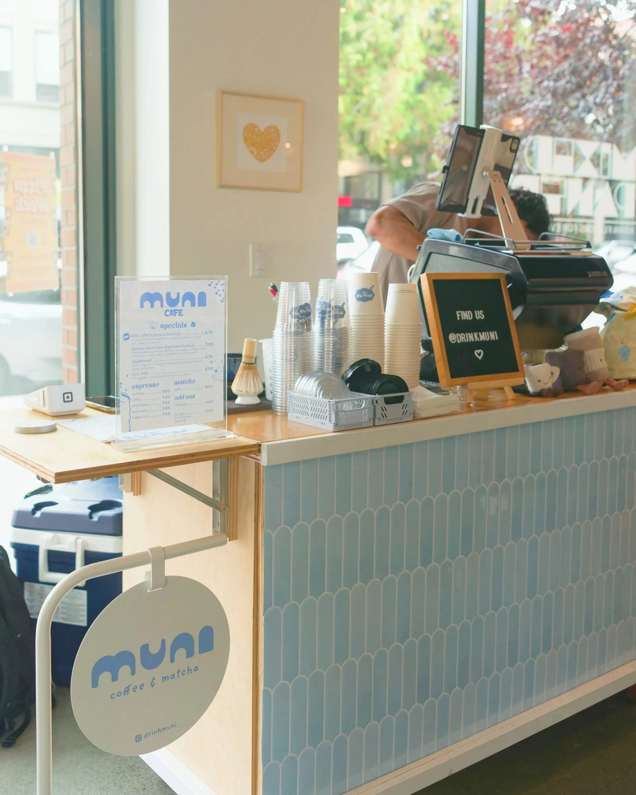 Coffee shop counter with cups, a menu, and a sign that says 'Find us @drinkmuni'. Large window showing outside greenery and a person working behind the counter.