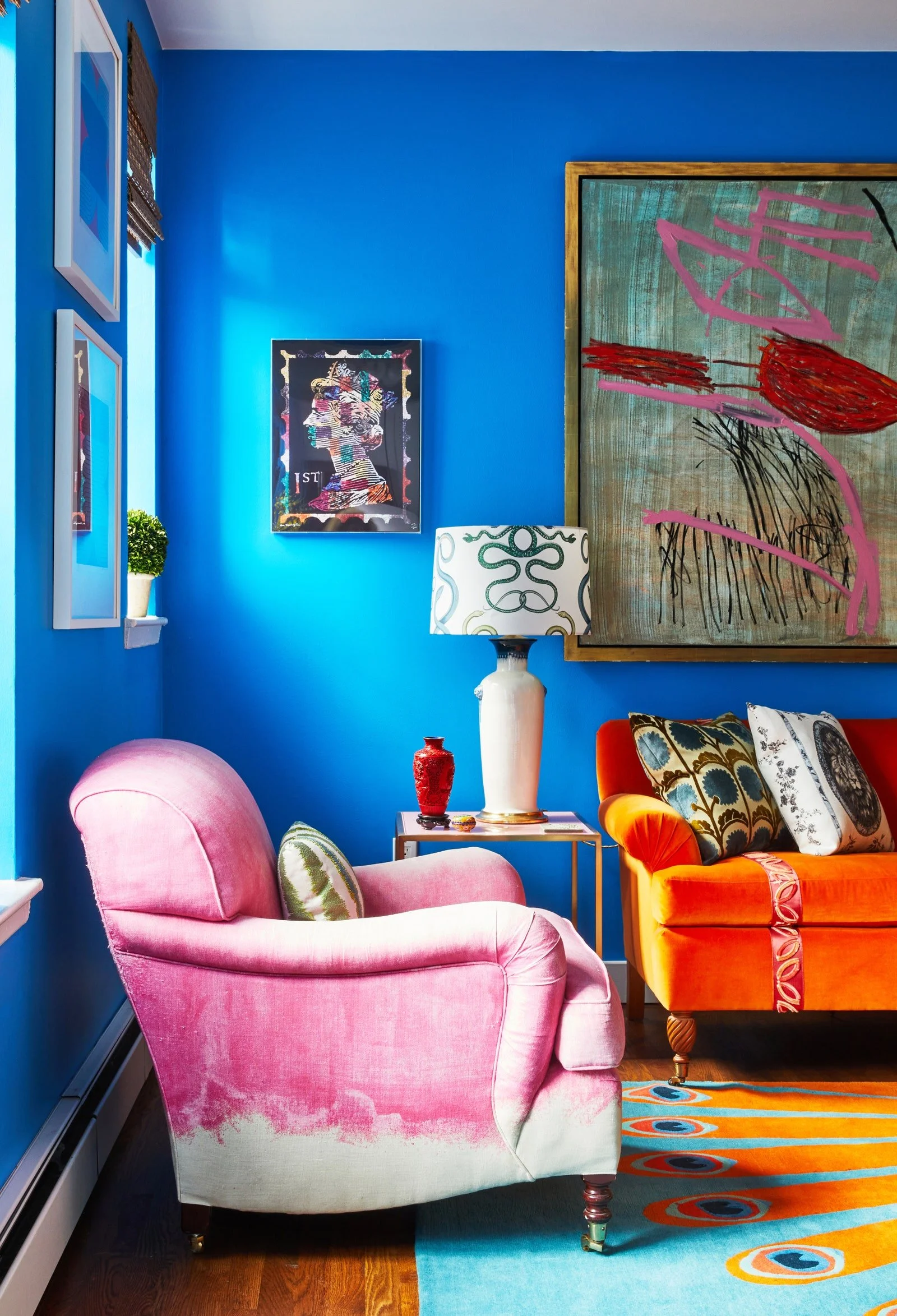 A vibrant living room with blue walls, a pink armchair, an orange couch with patterned pillows, a white table with a tall white lamp, and colorful abstract art on the walls.