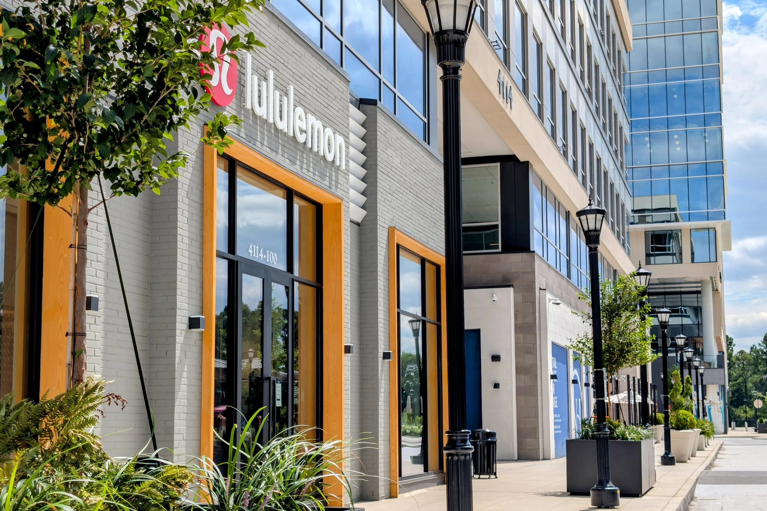 Modern commercial building with large glass windows, outdoor planters, and sidewalk lined with black street lamps