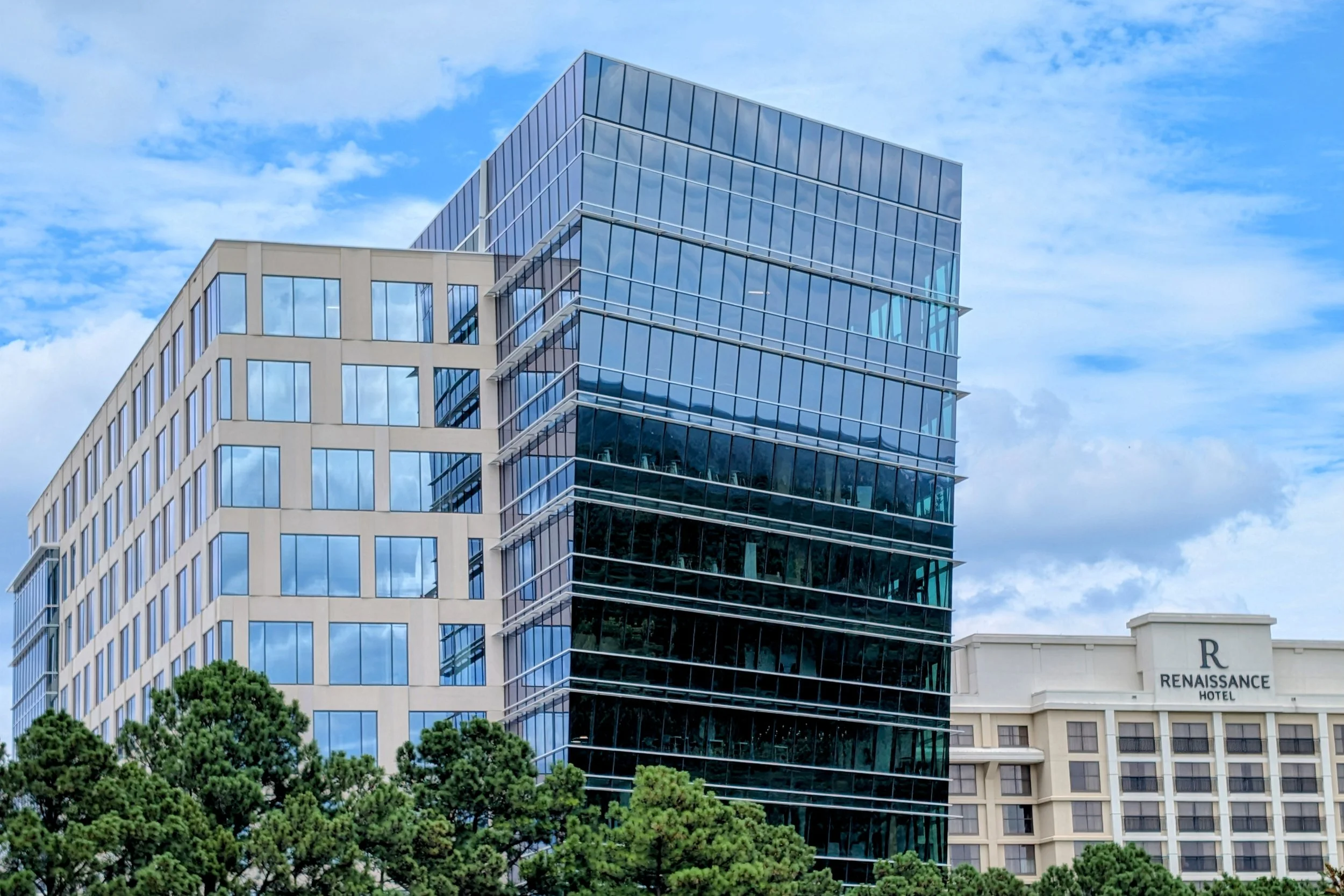 Modern office building with blue glass windows, adjacent to an older hotel labeled 'Renaissance Hotel,' with green trees in the foreground and a partly cloudy sky above.