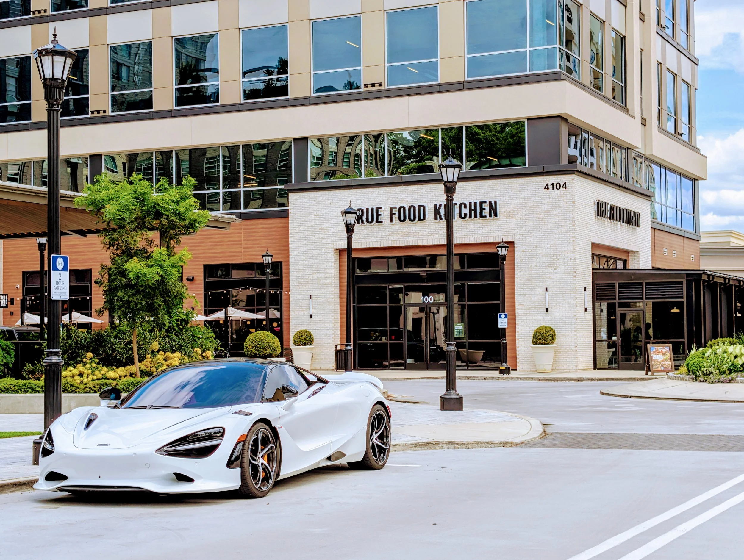 A white sports car parked on the street in front of a building with a restaurant called True Food Kitchen, modern glass and brick architecture, with small trees and street lamps nearby.