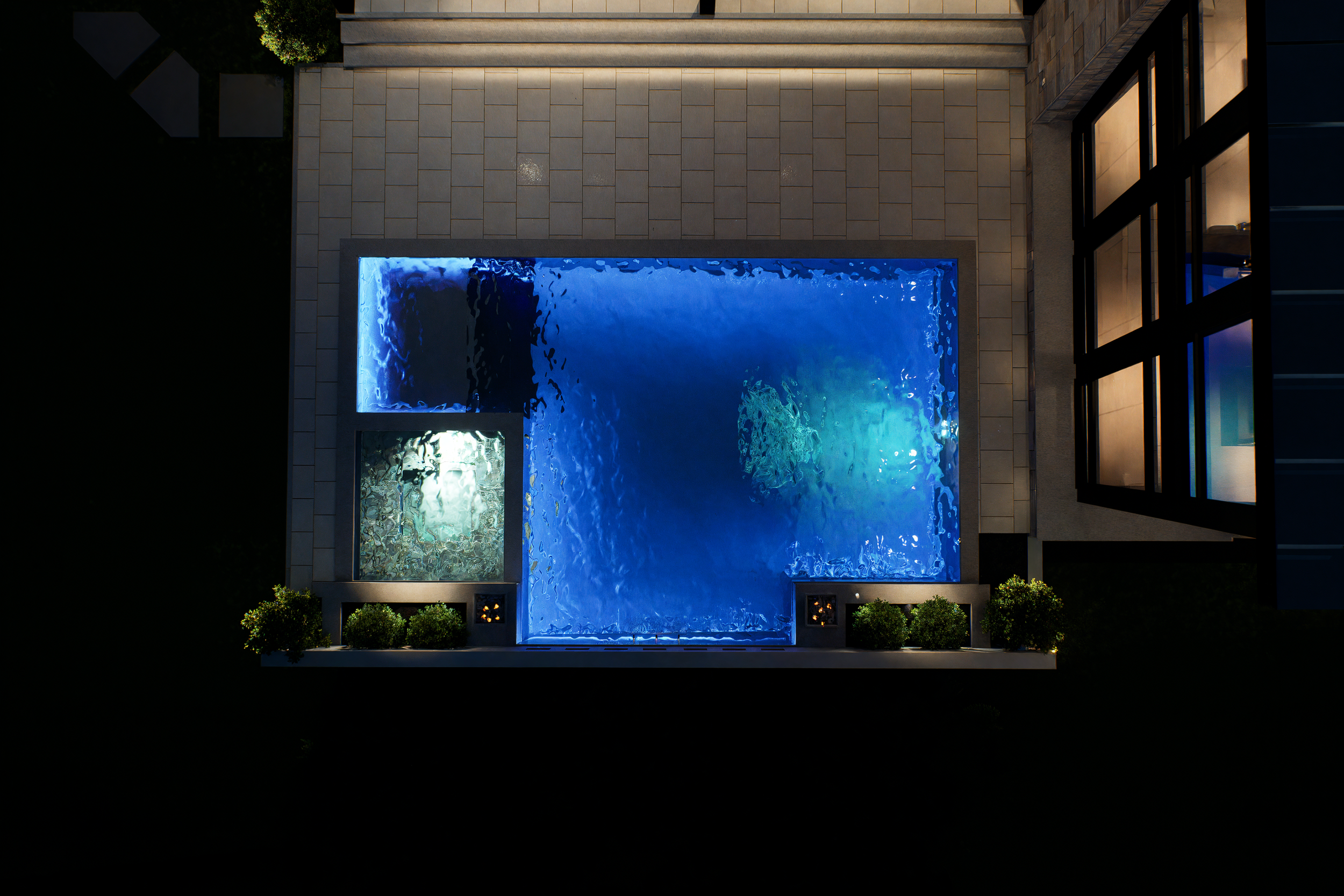 Top-down view of a house outdoor area at night with a lit rectangular swimming pool, a smaller square hot tub, surrounding paving, potted plants, and part of a modern building with large windows.