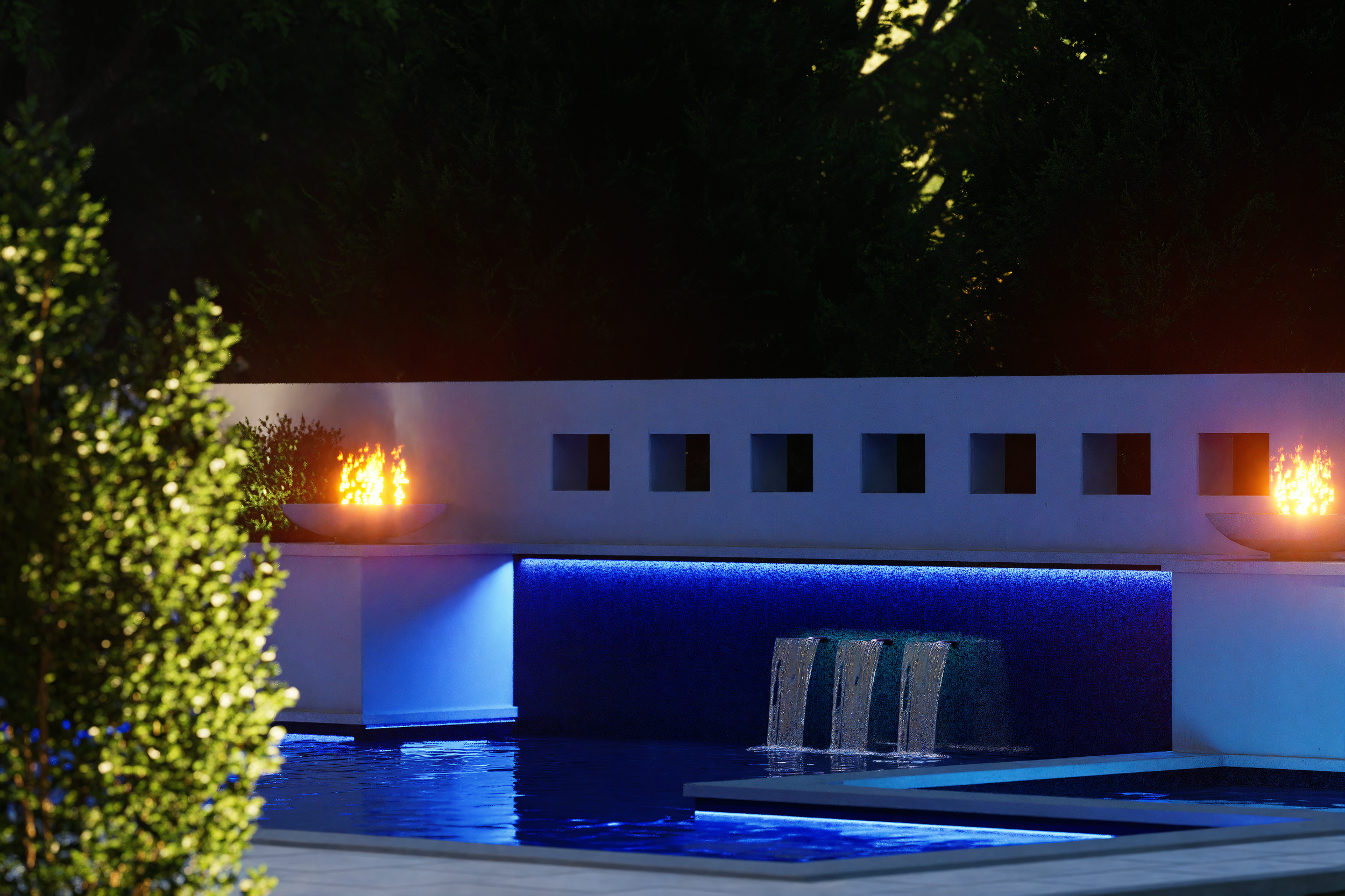Night view of a modern swimming pool with a water feature illuminated by blue lights, in front of a white wall with small square openings, topped with two lit fire bowls, surrounded by trees.
