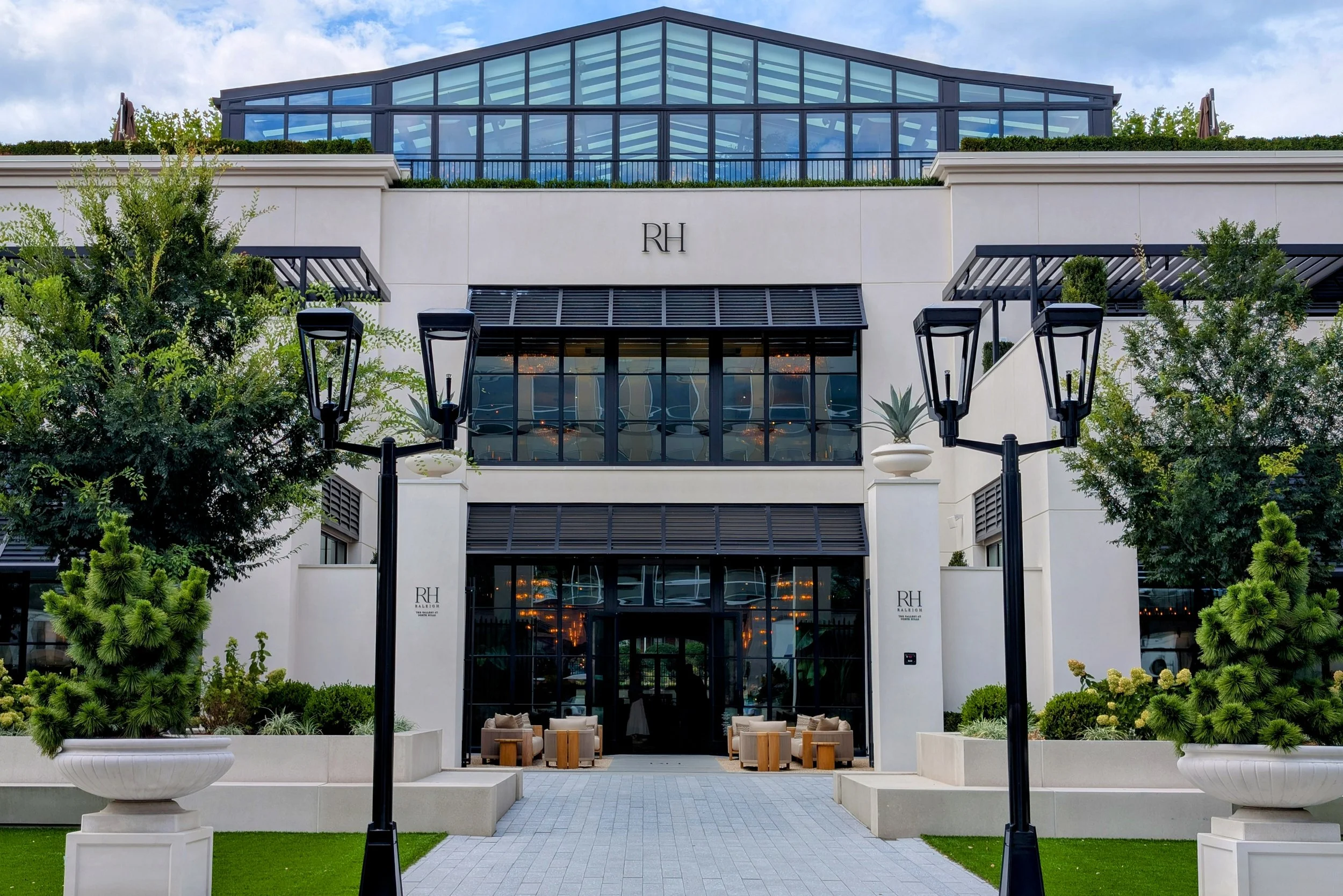 Modern multi-story hotel with glass facade, large potted plants, and outdoor seating area in front, surrounded by trees and decorative lamp posts.