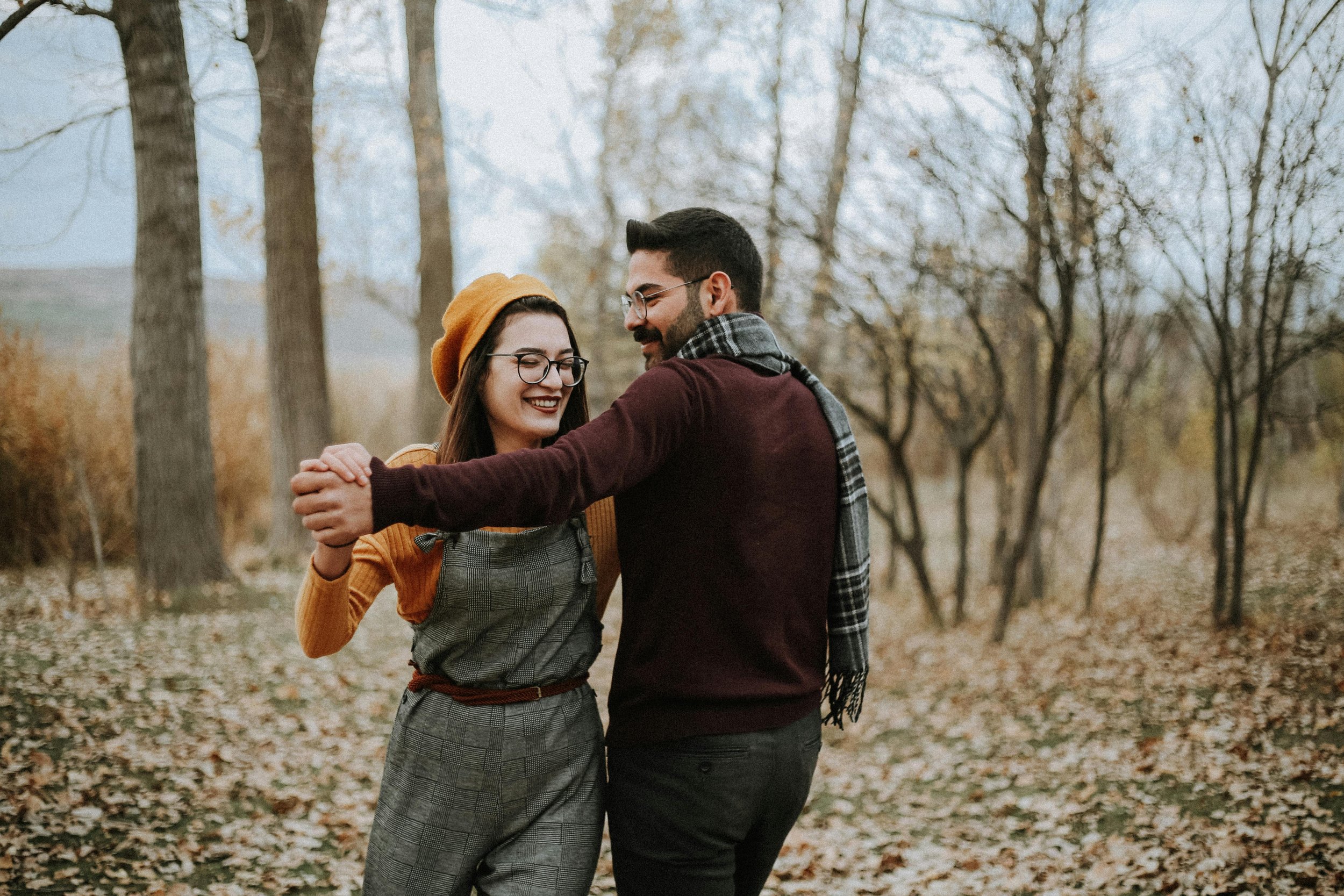 A young couple dancing and smiling outdoors in a wooded area during autumn, with fallen leaves on the ground.