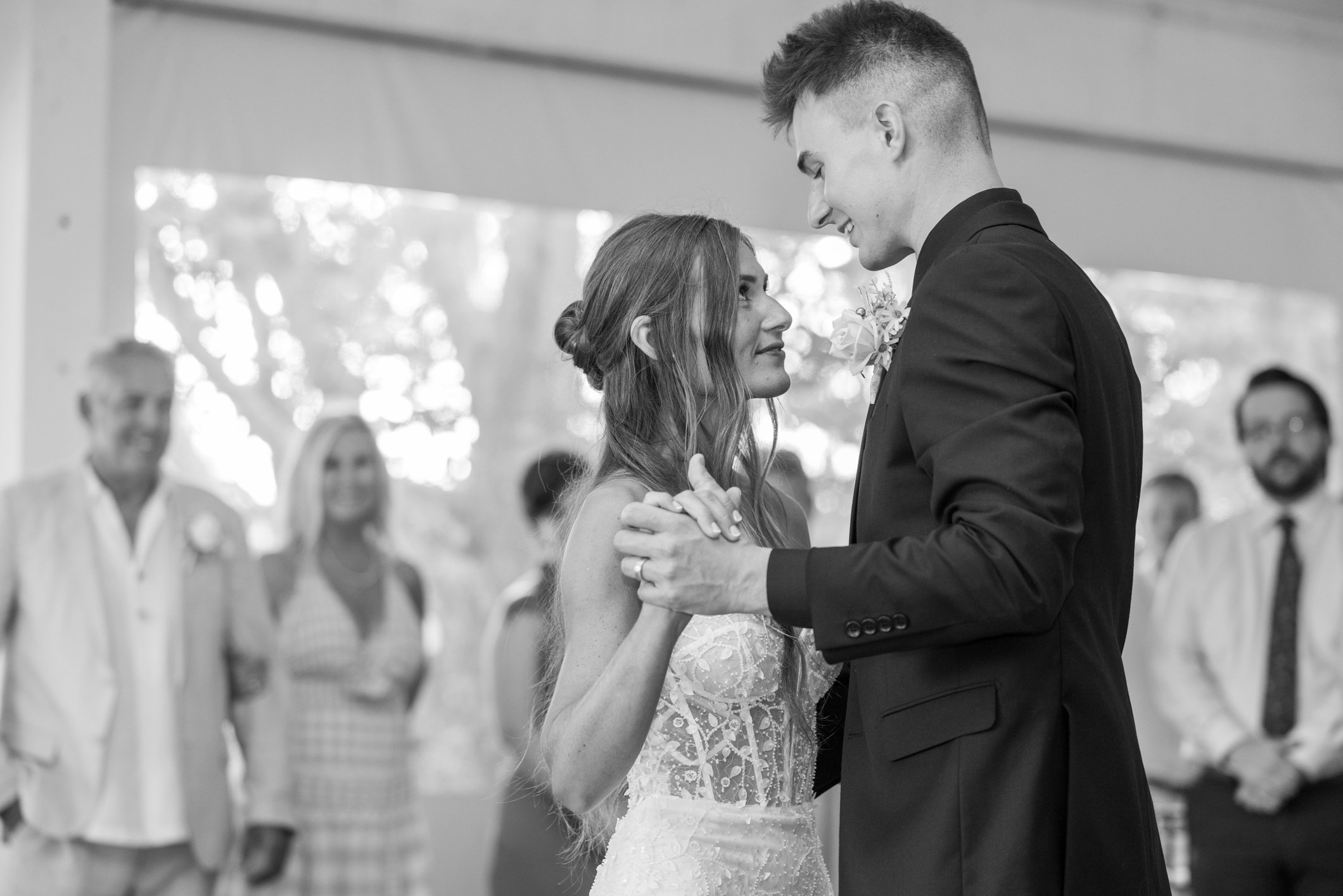 A bride and groom dancing closely during their wedding reception, gazing into each other's eyes while guests watch in the background.