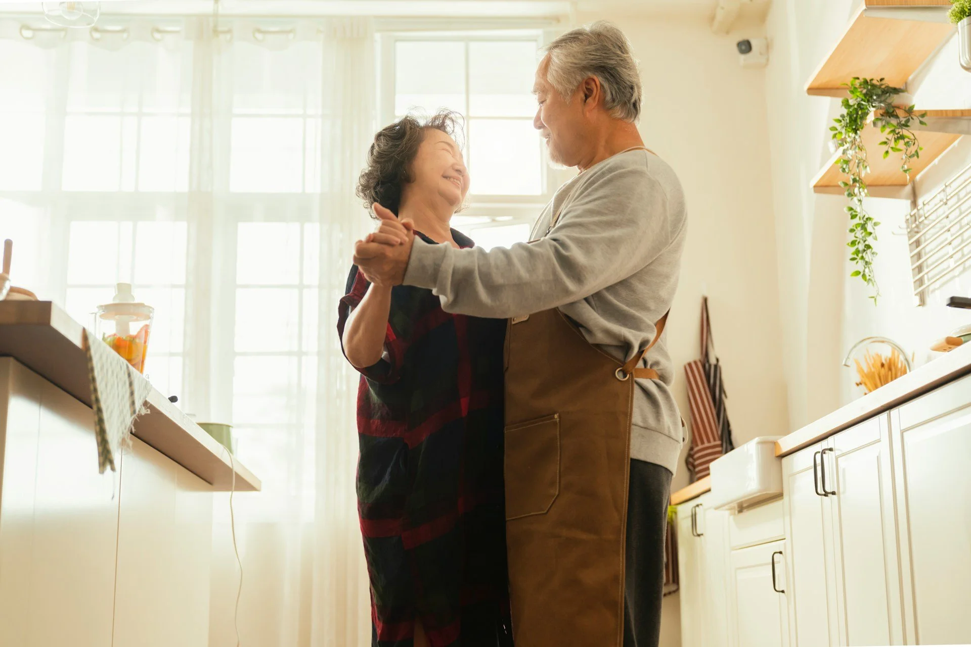 An elderly couple dancing and smiling together in a bright kitchen, holding hands.