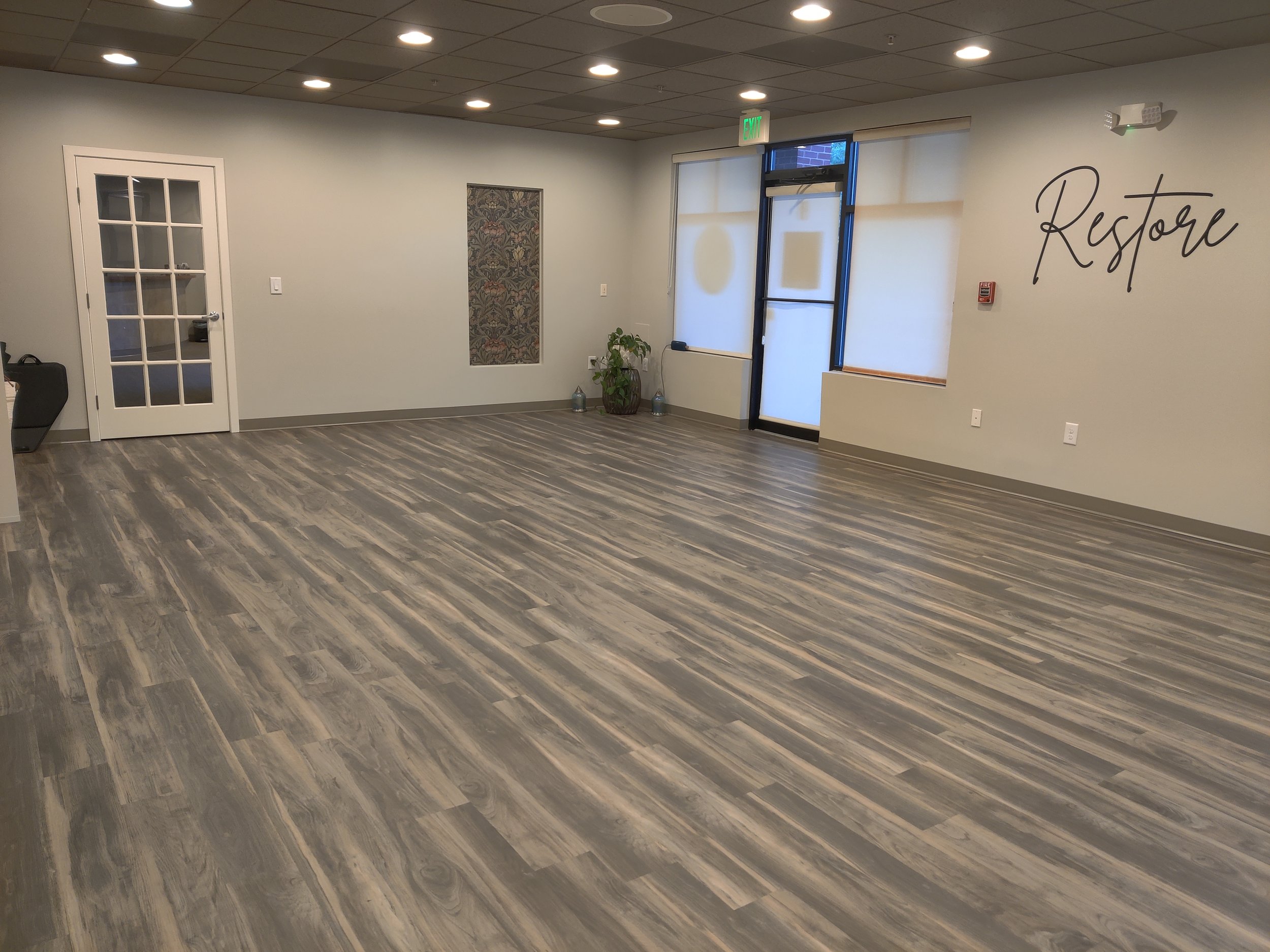 Indoor room with wood flooring, light walls, a door with glass panes, several large windows, a potted plant, wallpaper, and the word 'Restore' written on the wall.