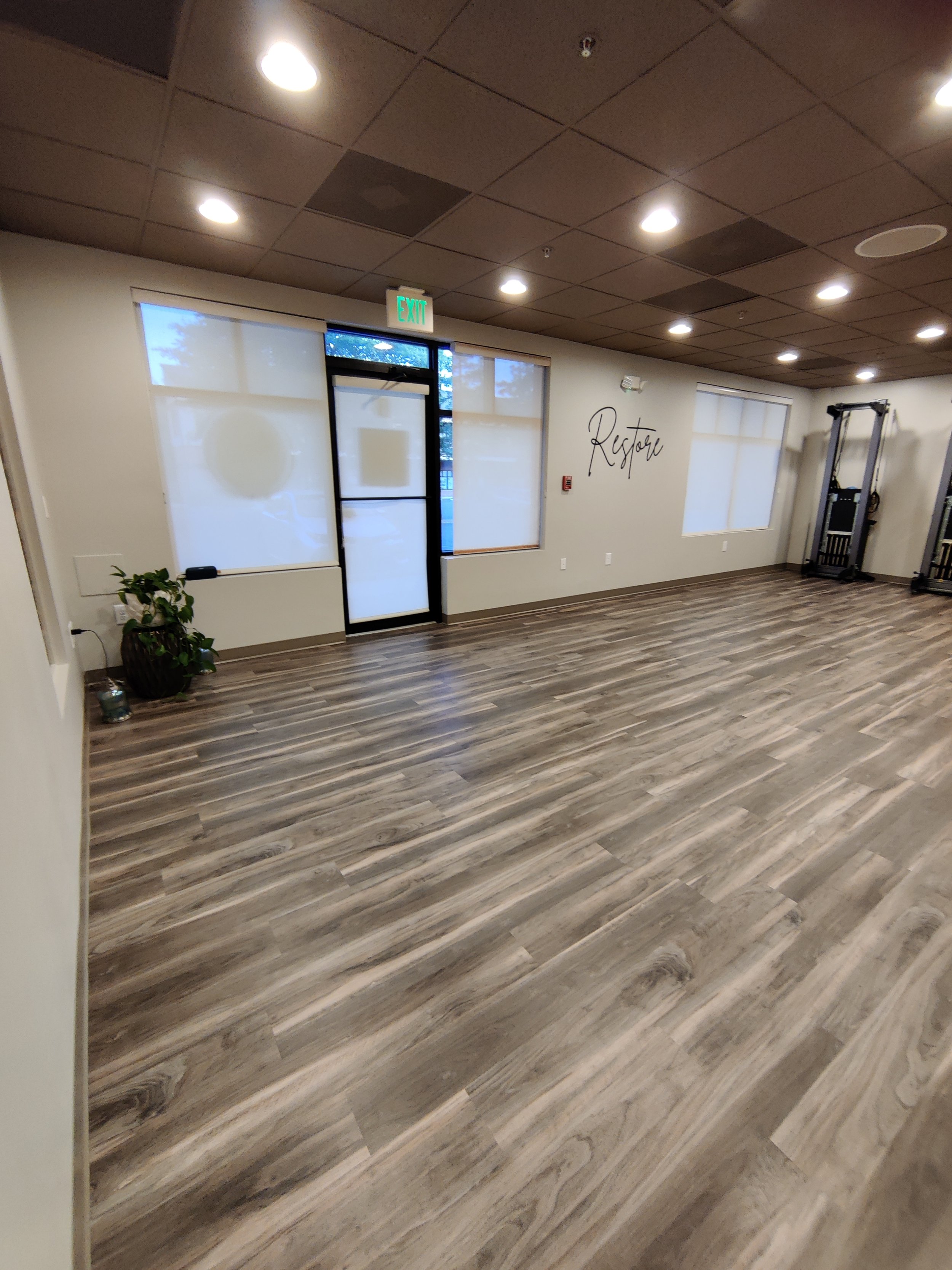 Interior of a wellness center studio with wood flooring, light walls, ceiling lights, a wall sign that reads 'Restore', and gym equipment against the wall.