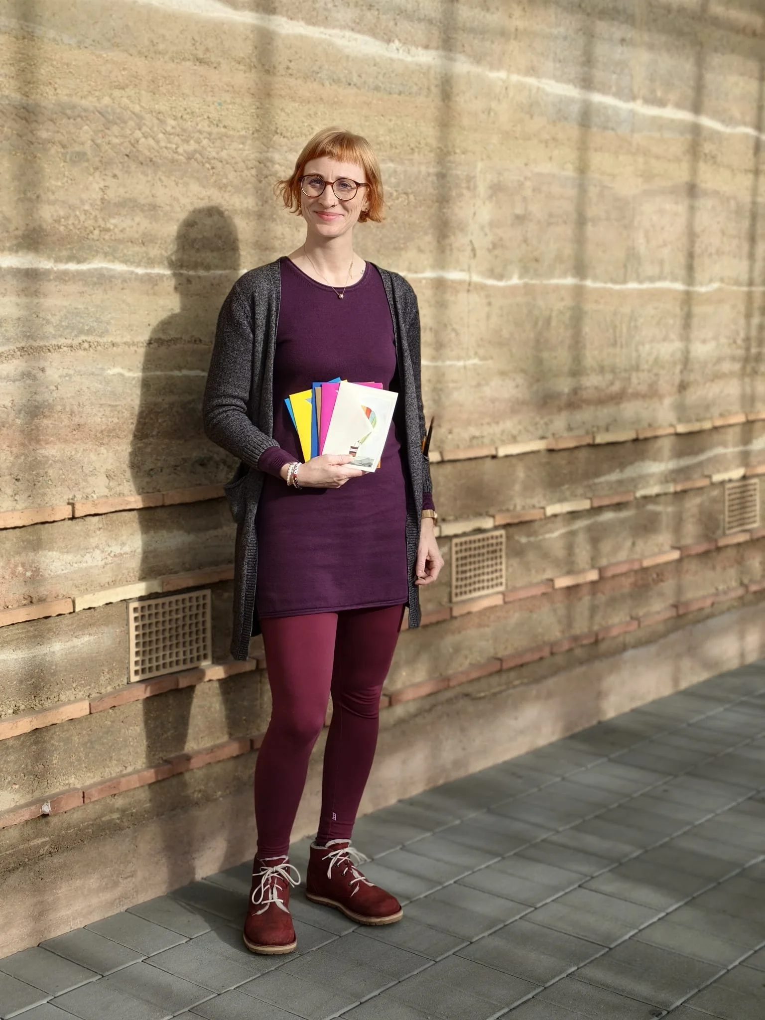 Eine junge Frau mit roten Haaren, Brille und einem Lächeln, die vor einer Steinmauer steht und mehrere bunte Bücher in den Händen hält.