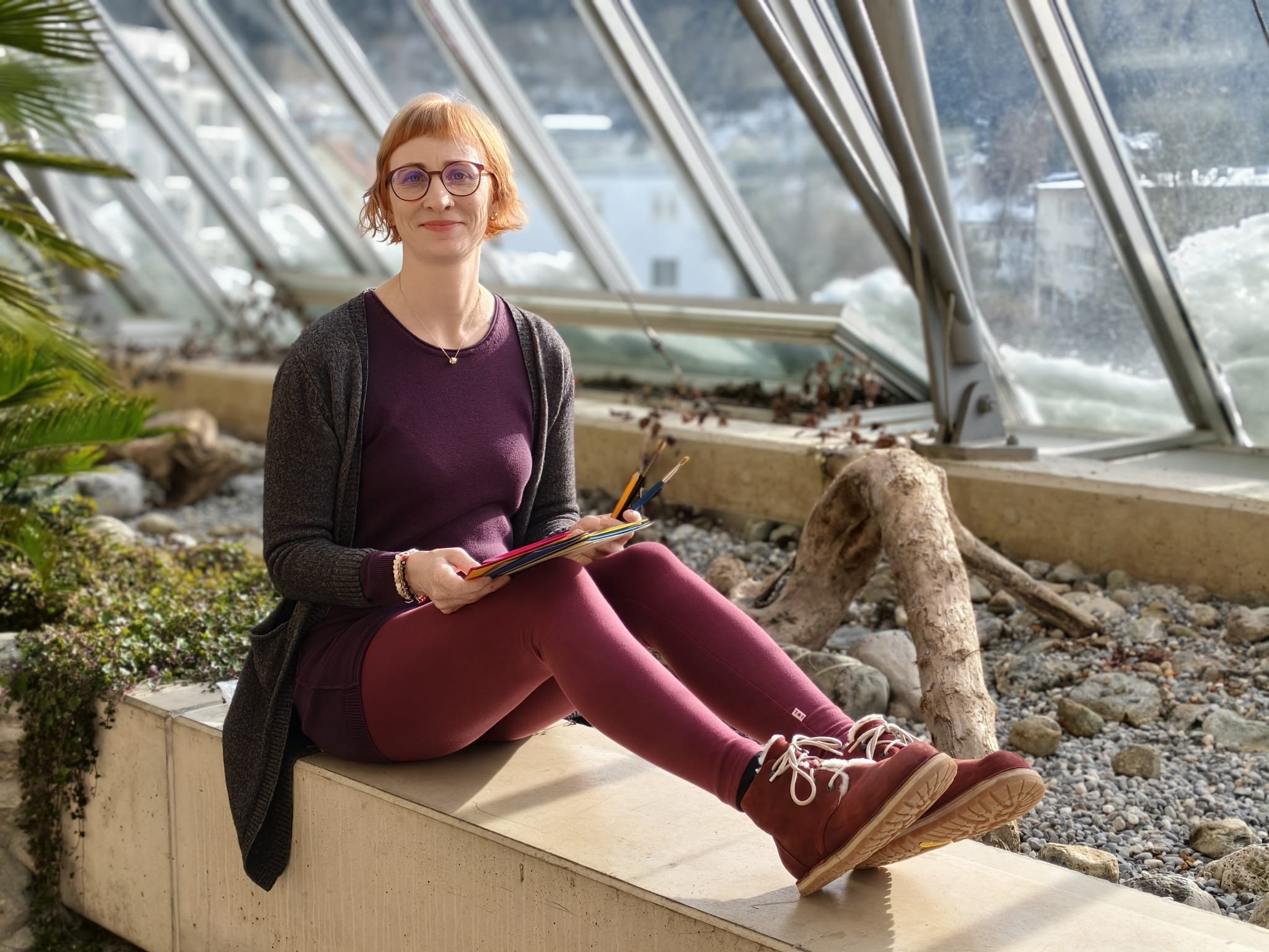 Eine Frau sitzt in einem Gewächshaus auf einer Betonkante, hält einen Stapel von farbigen Notizblättern und Blick in die Kamera. Im Hintergrund sind Fenster mit Blick auf verschneite Landschaft.