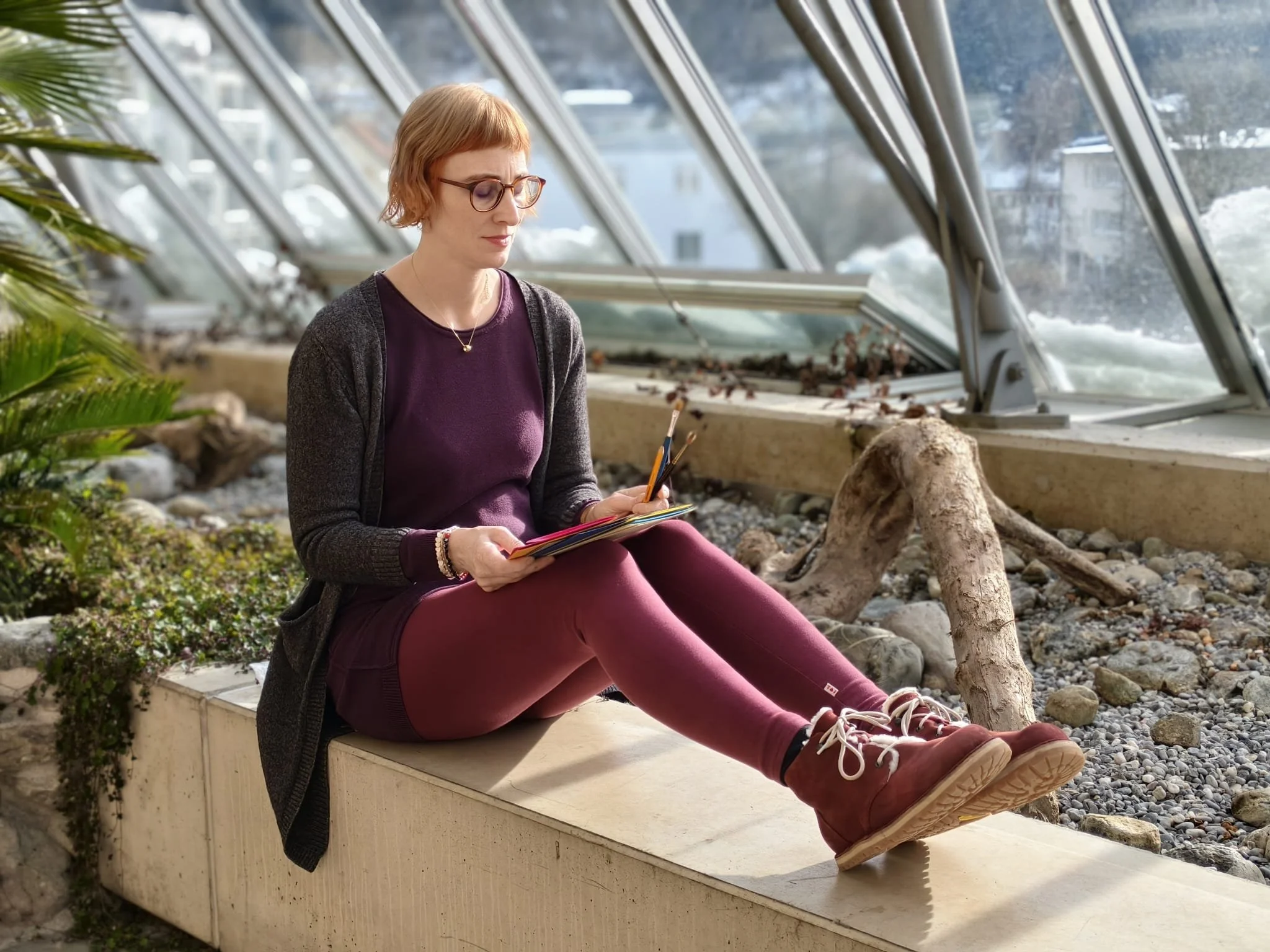 Eine Frau sitzt im Gewächshaus auf einem Betonsockel, liest ein Buch und hat einen Pinsel in der Hand. Sie trägt Brille, dunkelrotes Kleid, einen grauen Cardigan, braune Stiefel und Armbänder.