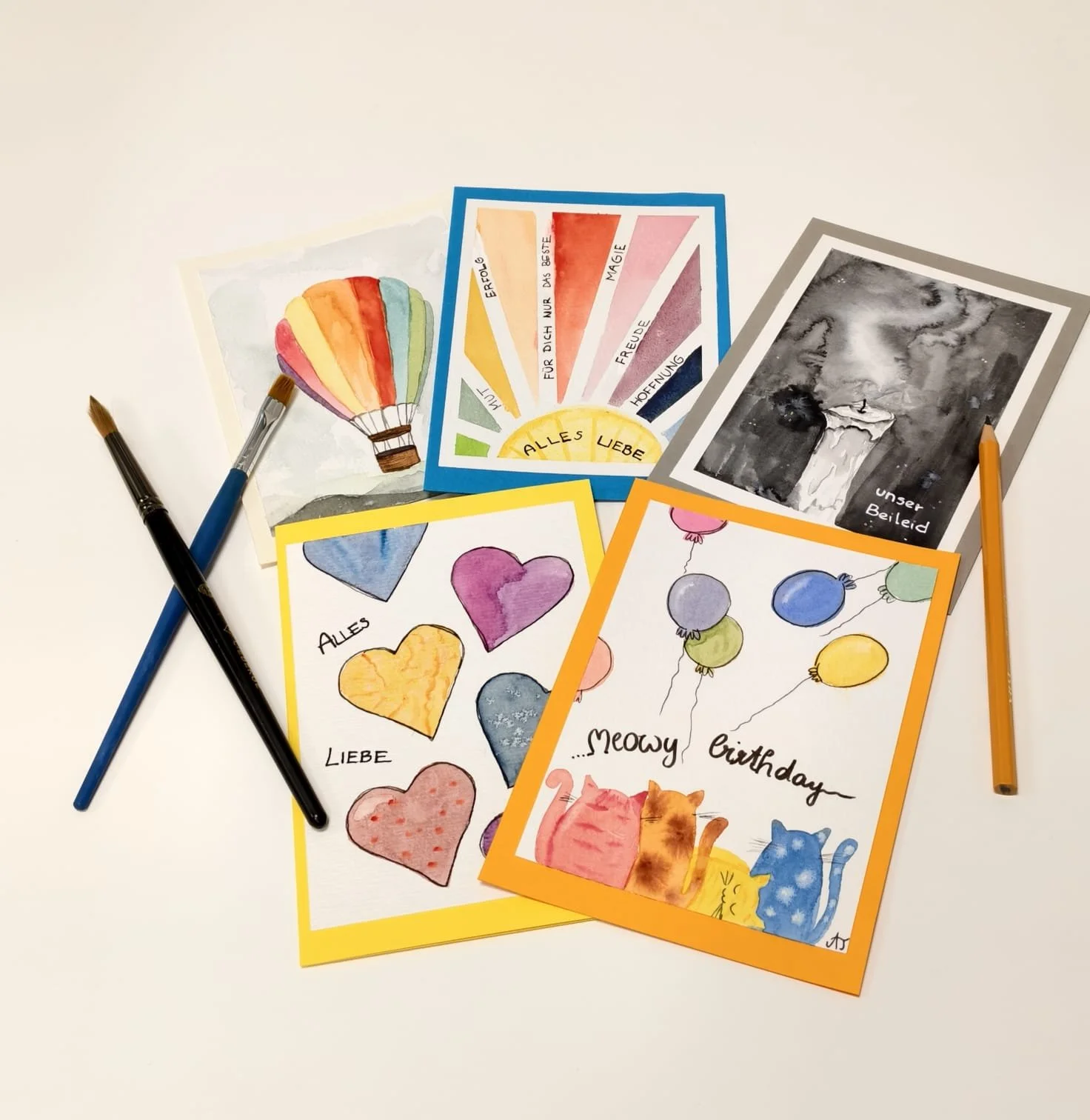 Verschiedene bunte Grußkarten mit Tiermotiven, Herzen, Ballons und Glückwunschtexten, umgeben von Malpinseln auf weißem Hintergrund.