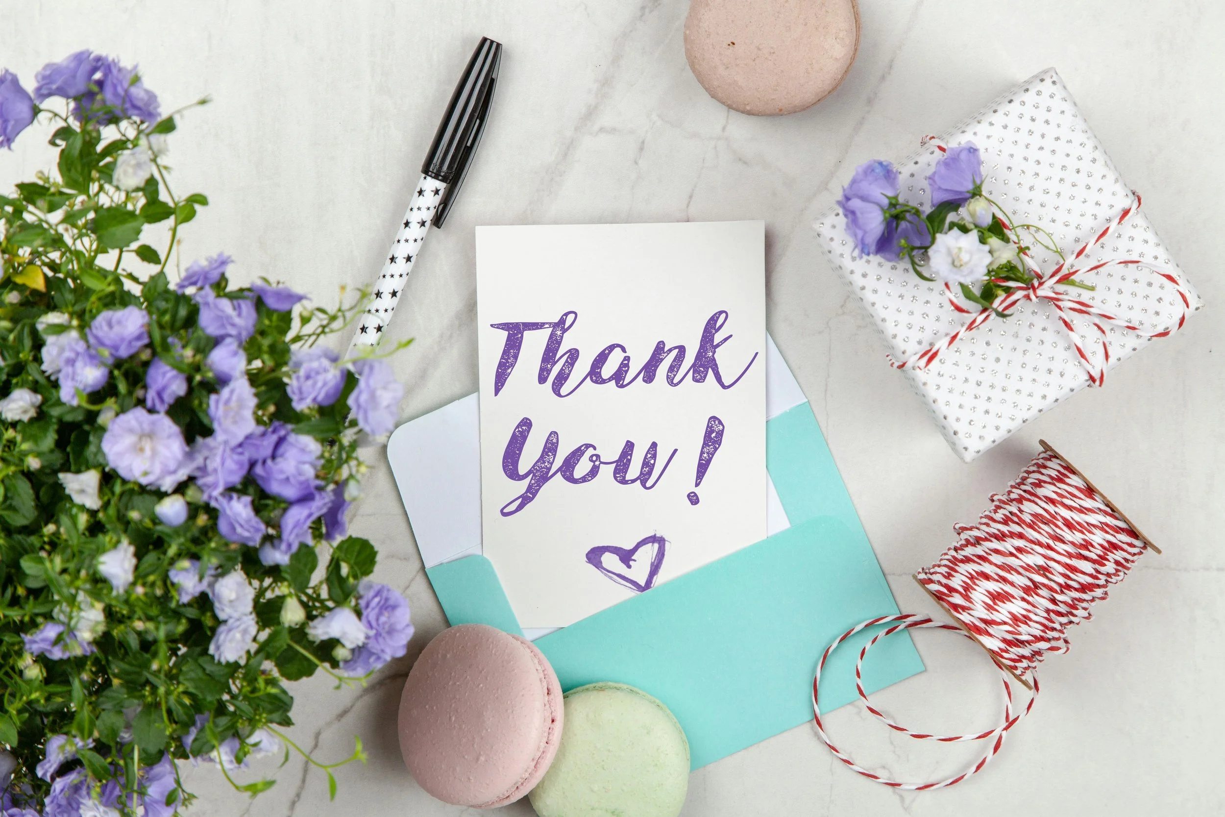 Blumen, eine Dankeskarte mit der Aufschrift 'Thank you!', Geschenkverpackung mit Blumen, Macarons, Schneidefaden, Kugel und Stift auf hellem Hintergrund.