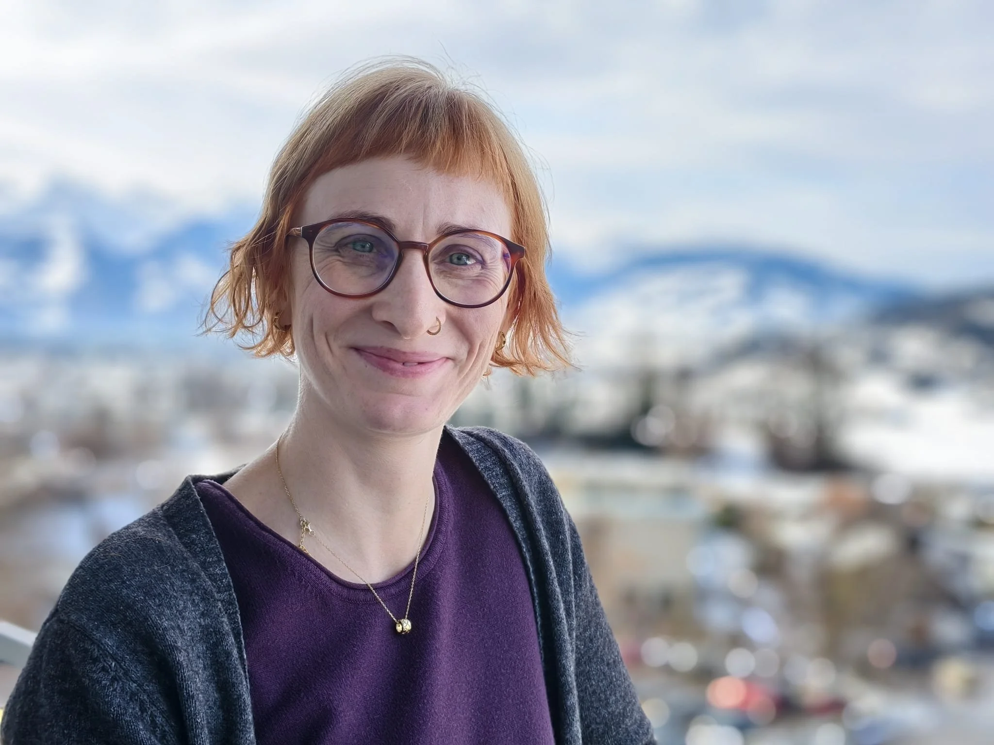 Porträt einer Frau mit roten Haaren, Brille, lila Oberteil und einer schwarzen Jacke vor einer Berglandschaft und Stadt im Hintergrund.