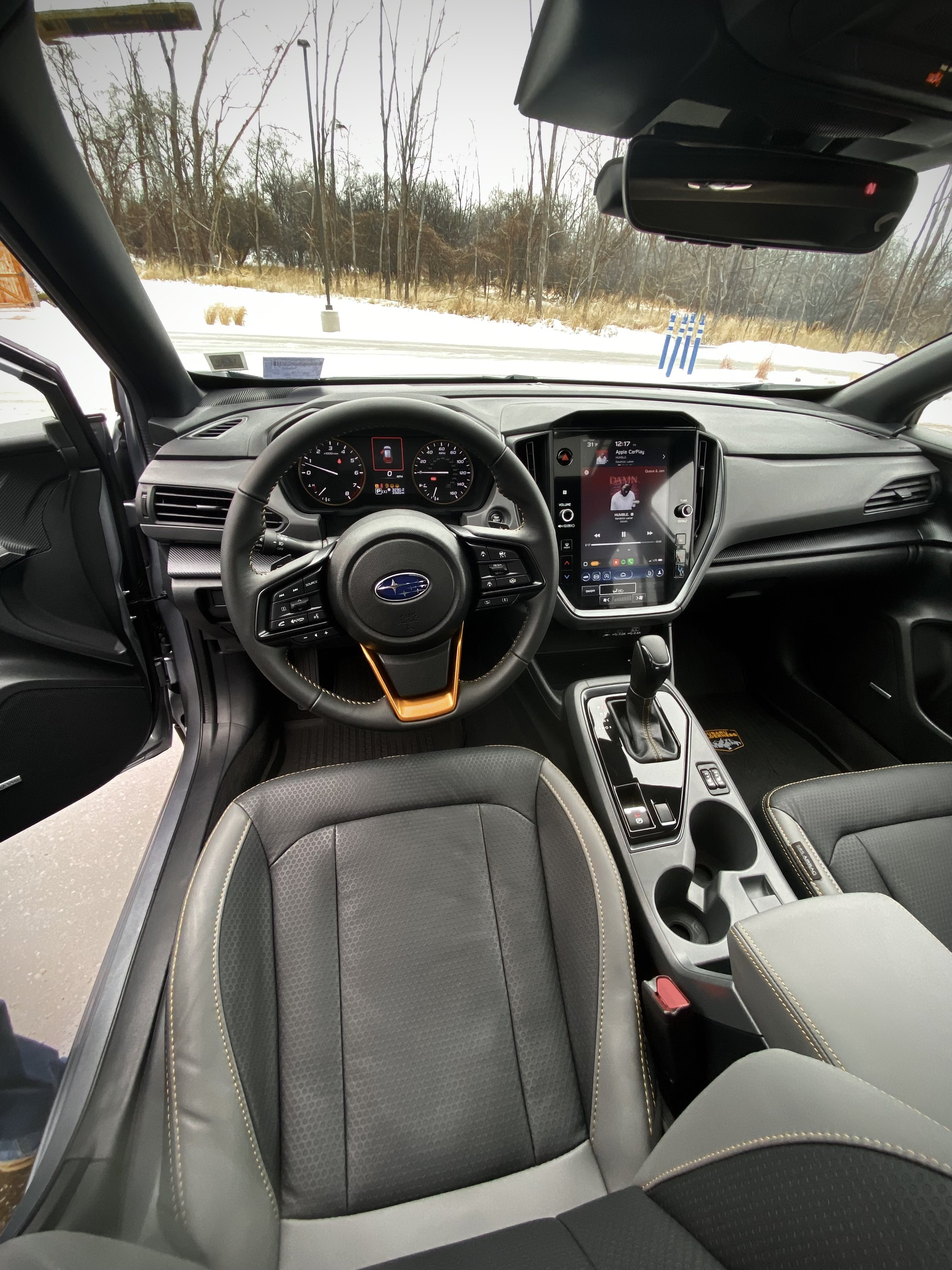 Interior view of a Subaru vehicle showing the dashboard, steering wheel, center console with gear shifter, and front seats, with snow and trees visible through the windshield outside.