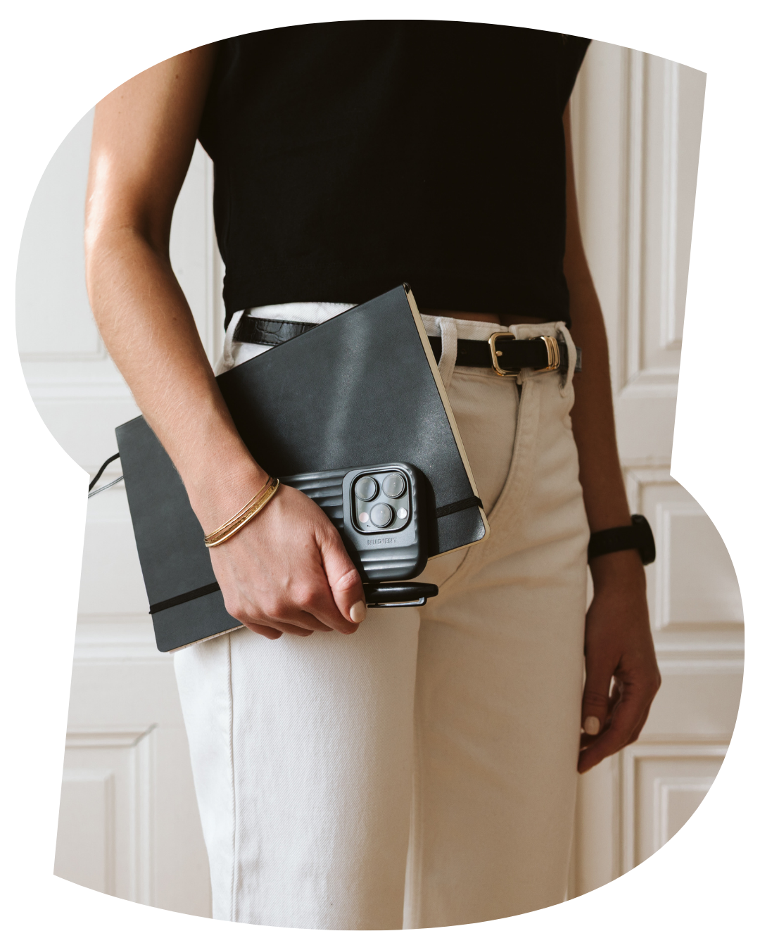 Eine Person trägt beige Hosen, ein schwarzes Oberteil und hält einen schwarzen Ordner, ein Smartphone und ein Notizbuch in der Hand.