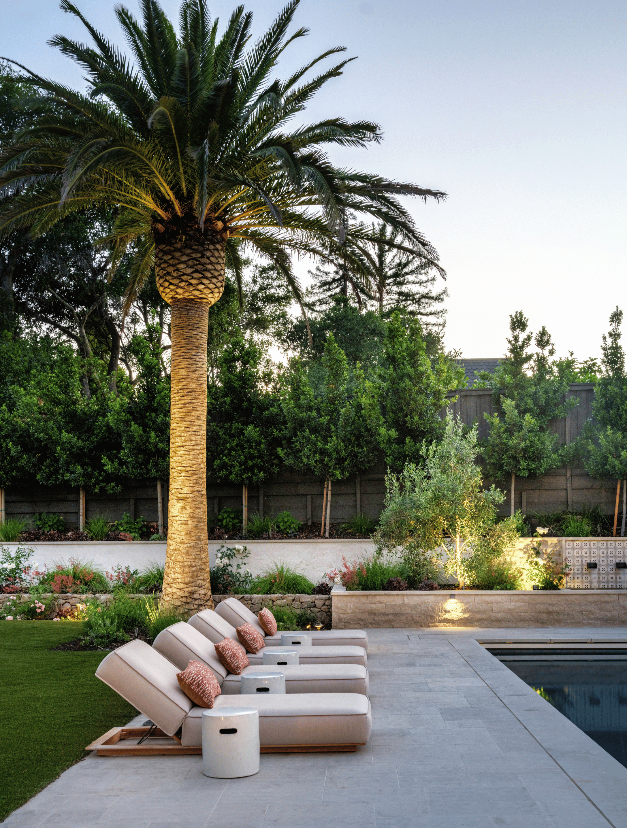 A backyard scene at dusk featuring a tall palm tree, a row of four white lounge chairs with pink pillows, a small white round side table, a grassy area, and a pool, all illuminated by outdoor lights.