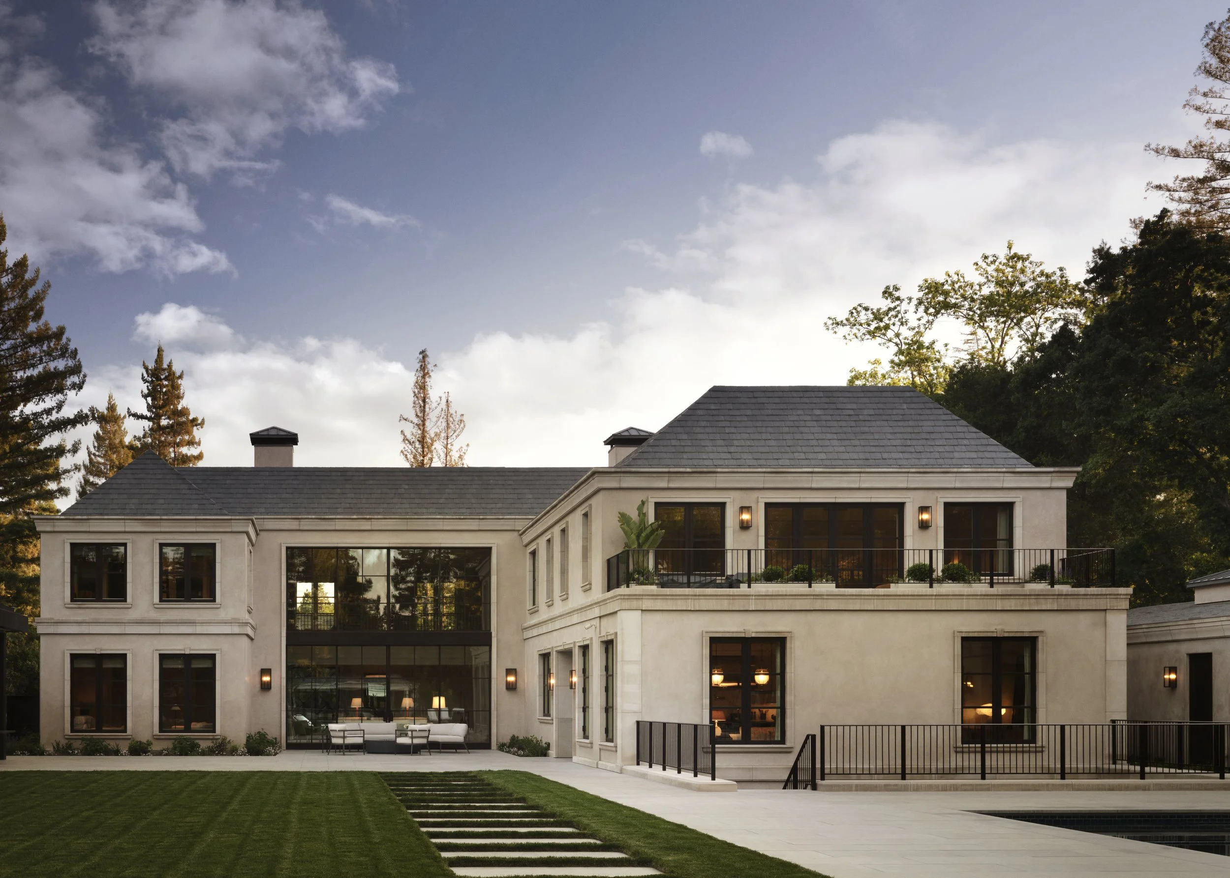Modern two-story house with large windows, balconies, and outdoor seating, surrounded by a well-manicured lawn and trees, under a partly cloudy sky.