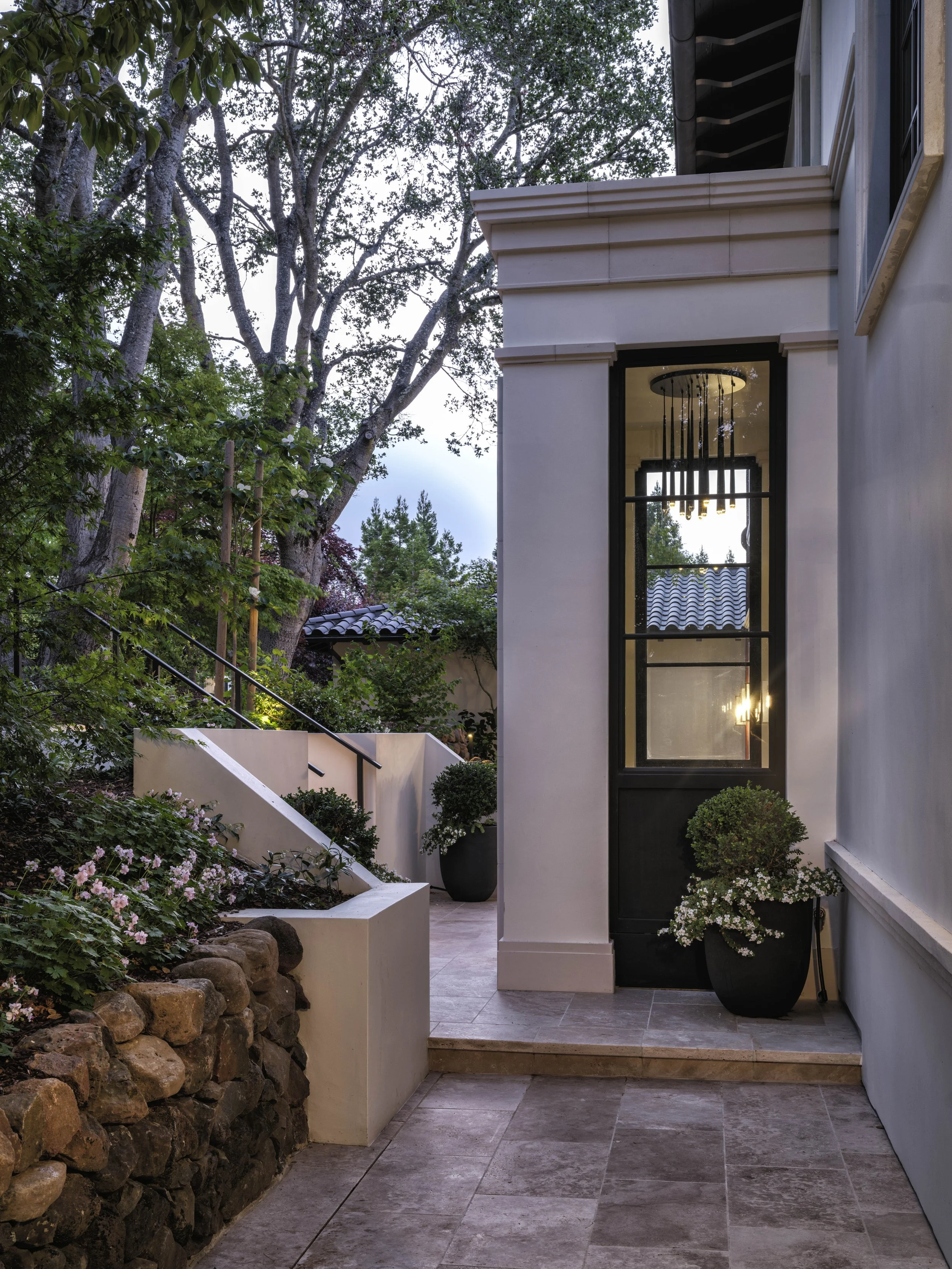 Elegant outdoor entrance with a pathway, stone wall, greenery, potted flowering plants, and a window with a chandelier visible inside.