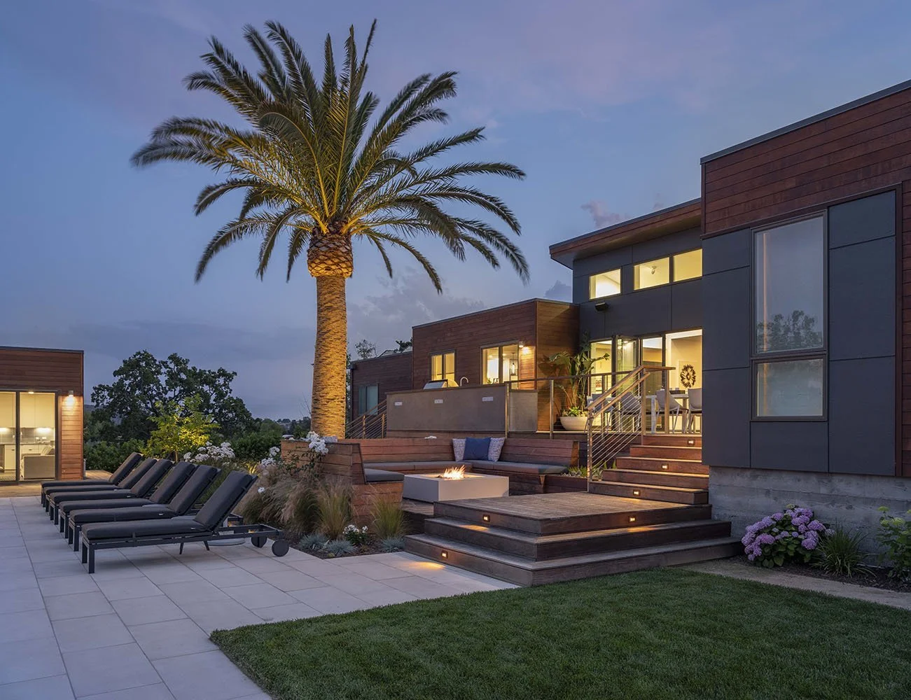 Modern residential backyard at dusk with a tall palm tree, outdoor lounge chairs, a fire pit, and a multi-story house with large windows and outdoor stairs, surrounded by landscaped garden and trees.