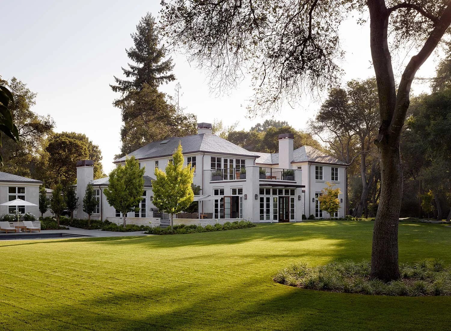 Large white house with multiple windows and a gray roof, surrounded by green lawn and trees in a spacious yard.