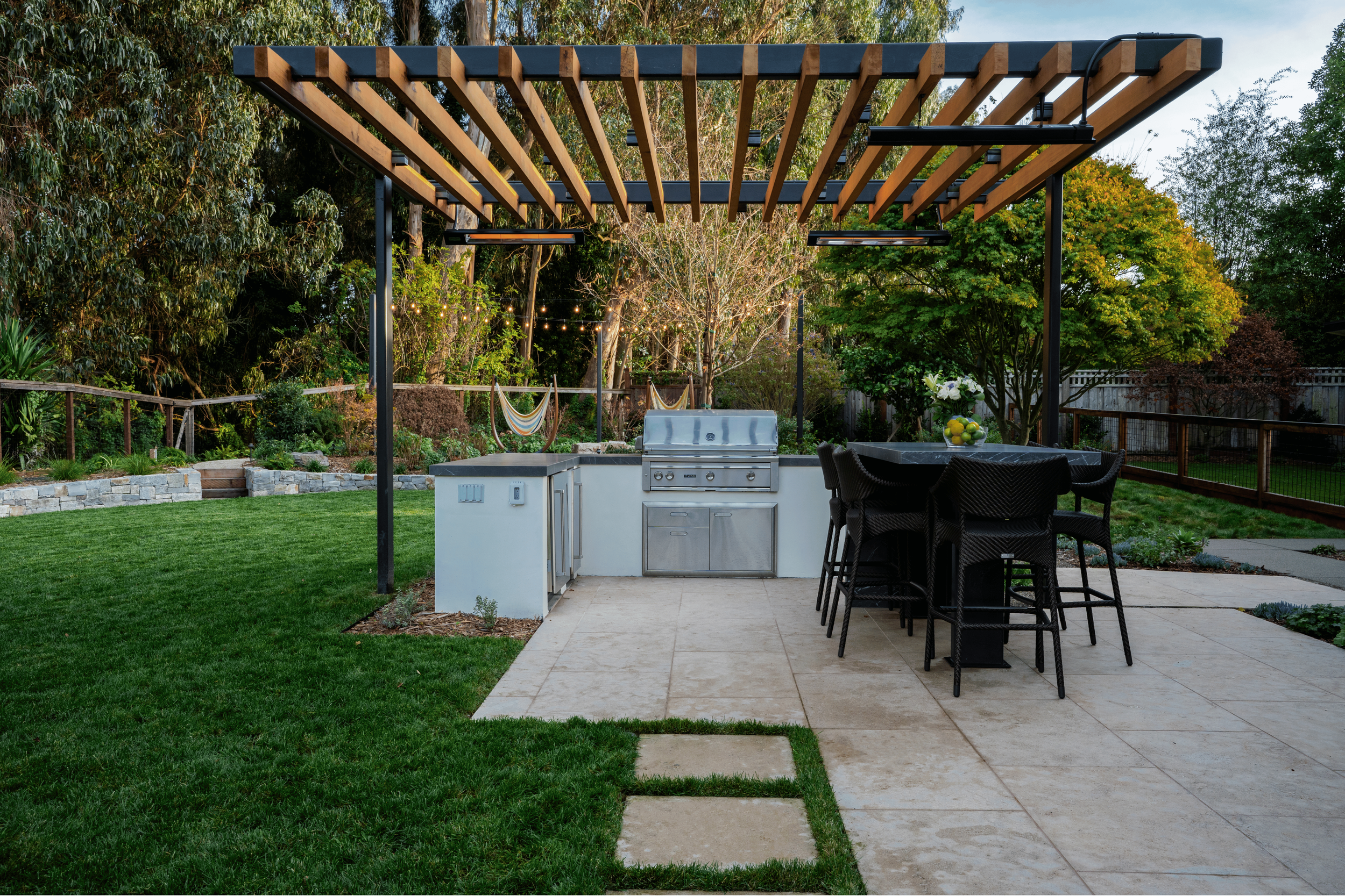 An outdoor backyard patio with a covered kitchen area that includes a built-in grill, counter space, and a table with six black chairs. There's a grassy lawn, a stone pathway, and trees with string lights in the background.