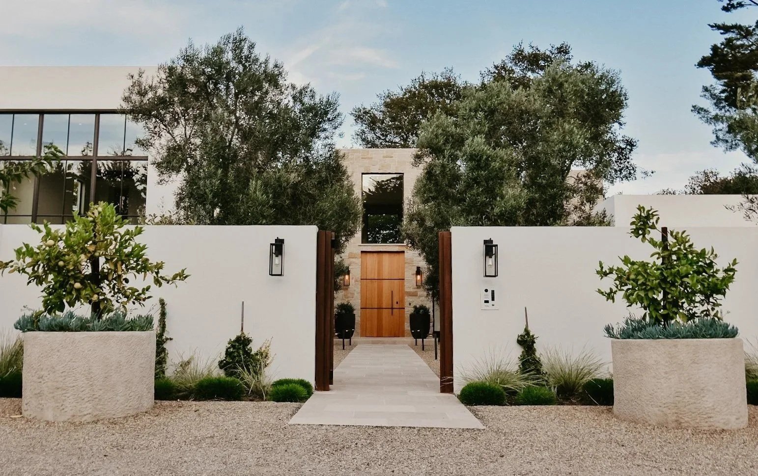 Modern house entrance with white walls, wooden door, potted plants, and trees surrounding the walkway.