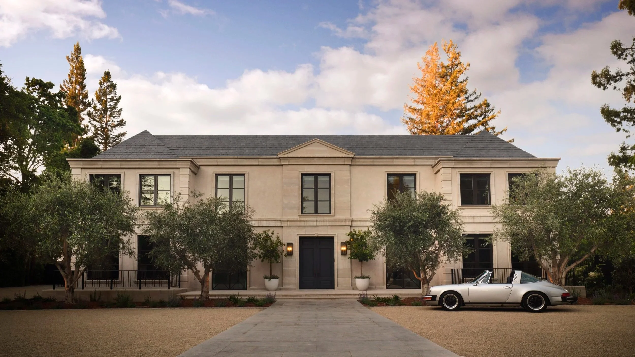 A two-story mansion with beige stone exterior, black window frames, and a central entrance flanked by two trees. A white convertible car is parked in the driveway on the right side of the house. The sky is partly cloudy with orange trees in the backg